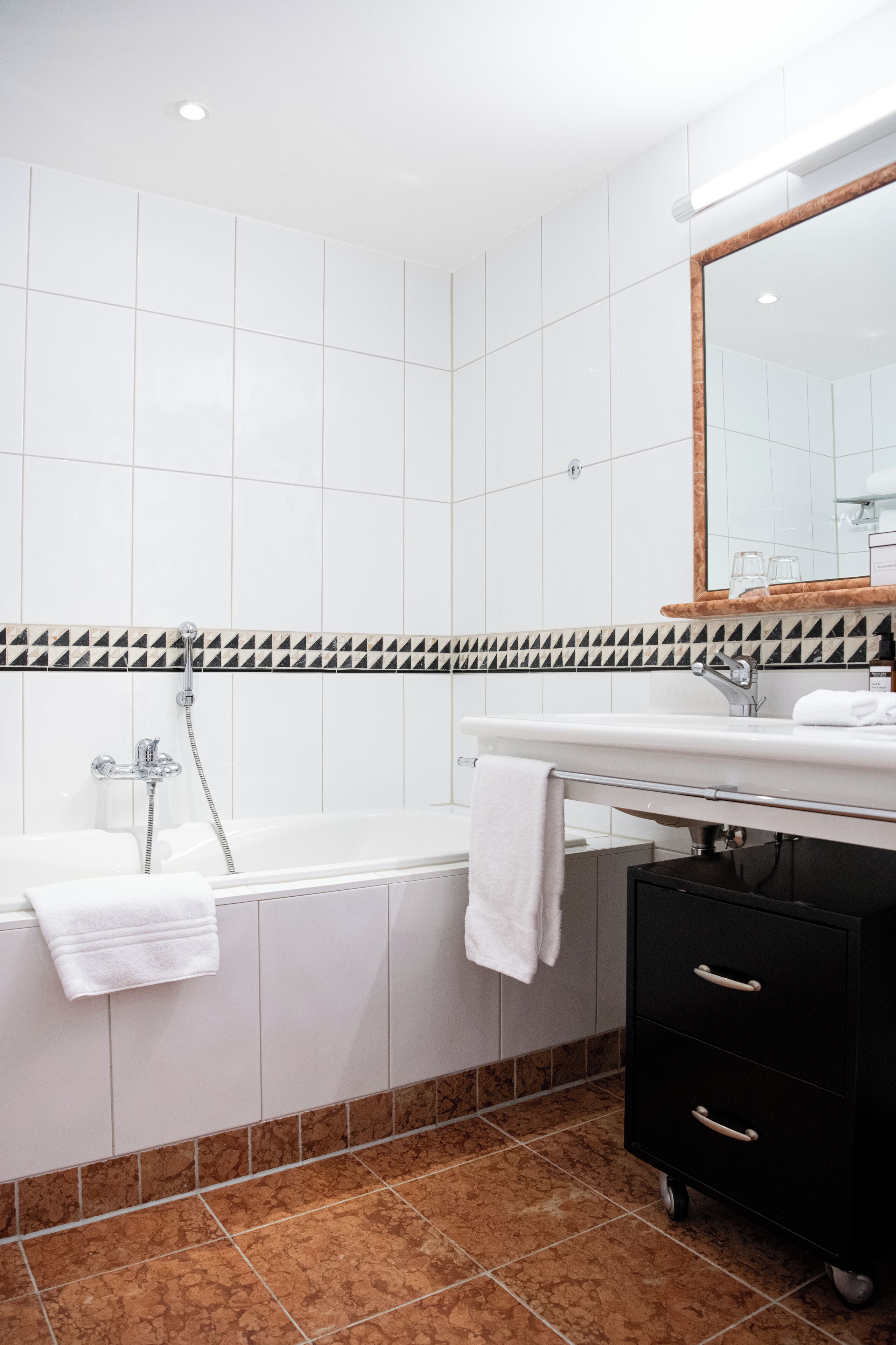 Een moderne badkamer met witte tegels, een zwart-wit accentstrook, een wit bad met een handdoek, een zwart wastafelmeubel met lades en een grote spiegel.