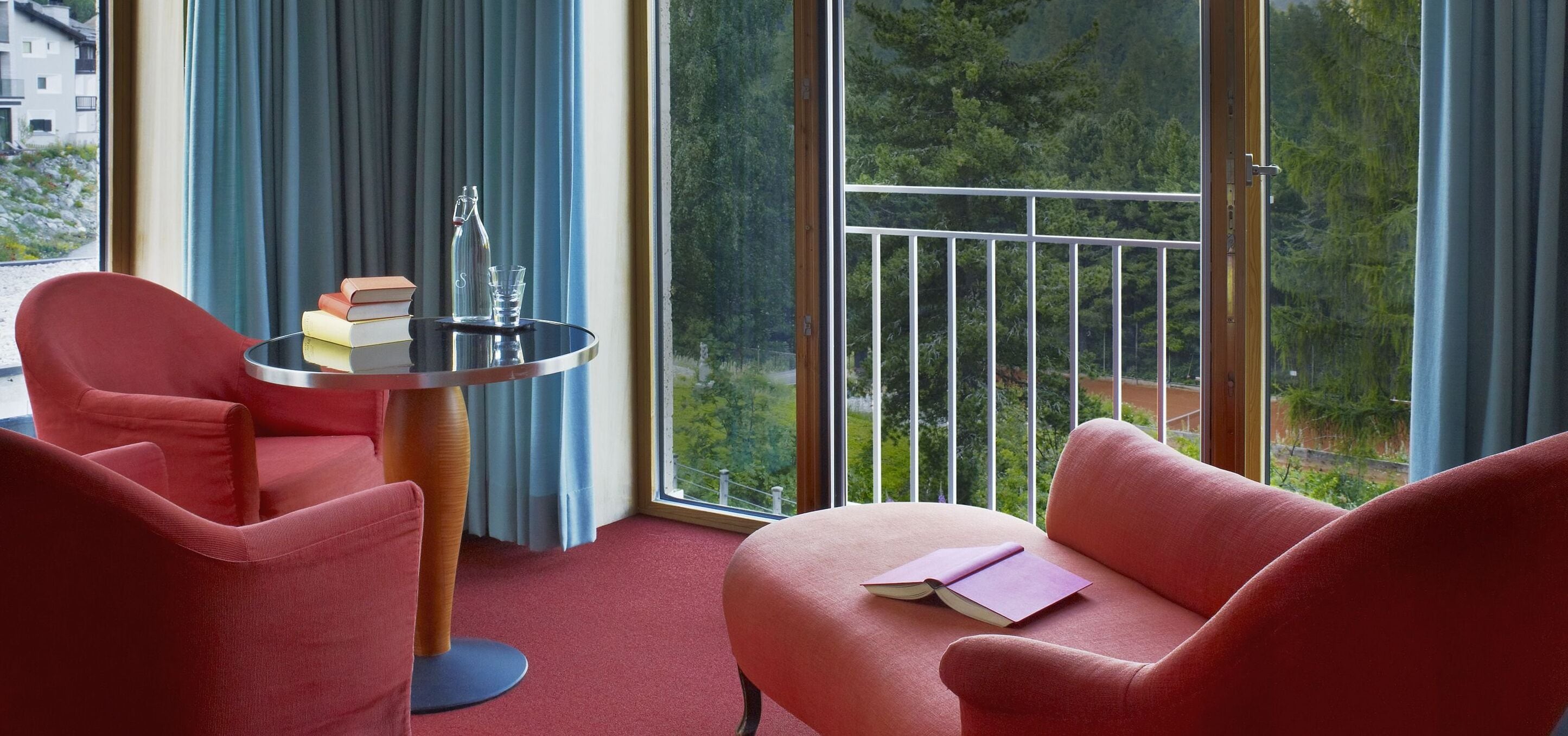Une pièce avec une moquette rouge, deux fauteuils rouges, une petite table ronde avec des livres et un verre, et un balcon doté d’une rambarde en métal et de plantes à l’extérieur.