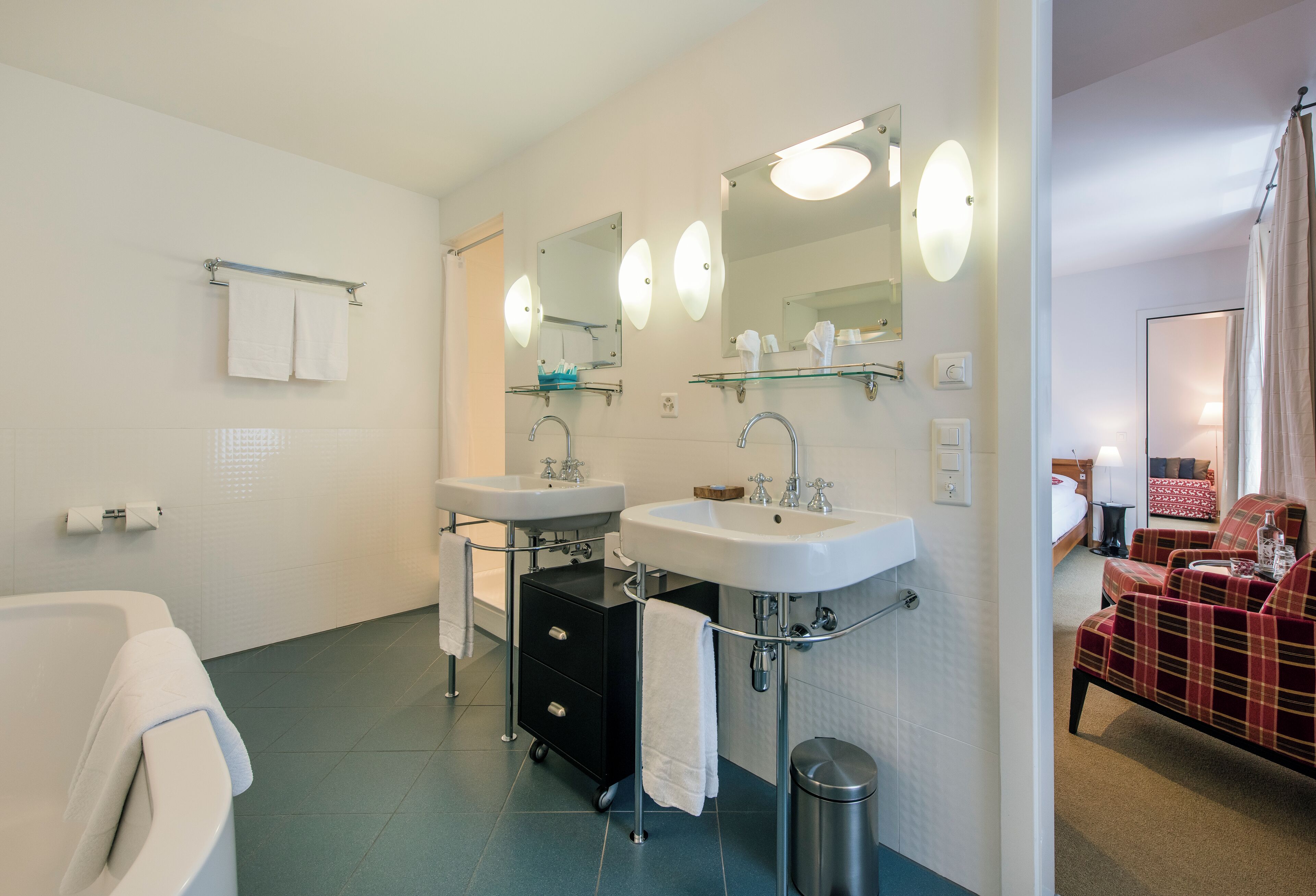 Un baño moderno con dos lavamanos, una bañera y un espejo con iluminación. Hay un cubo de basura y un perchero para toallas.