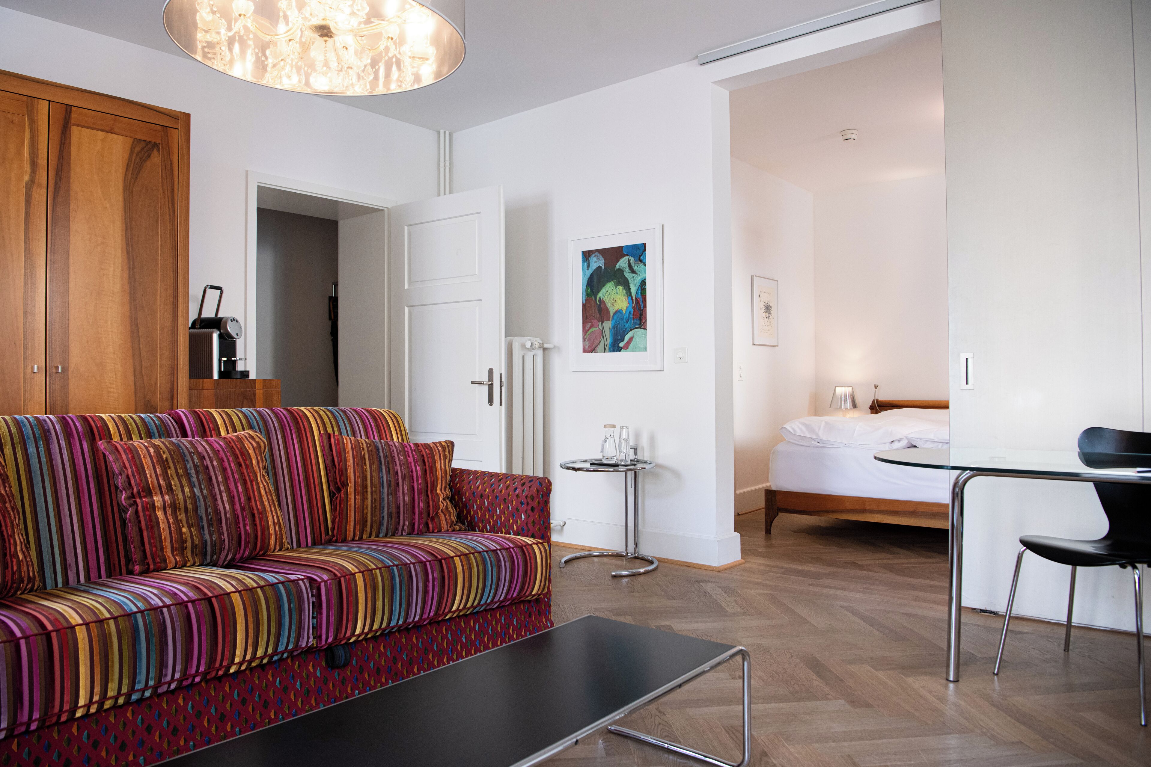 A modern living room with a colorful sofa, a small table, and a bed in the background.