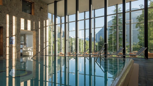 Una piscina interior con paredes de vidrio transparente, sillones de descanso y vistas a las montañas.