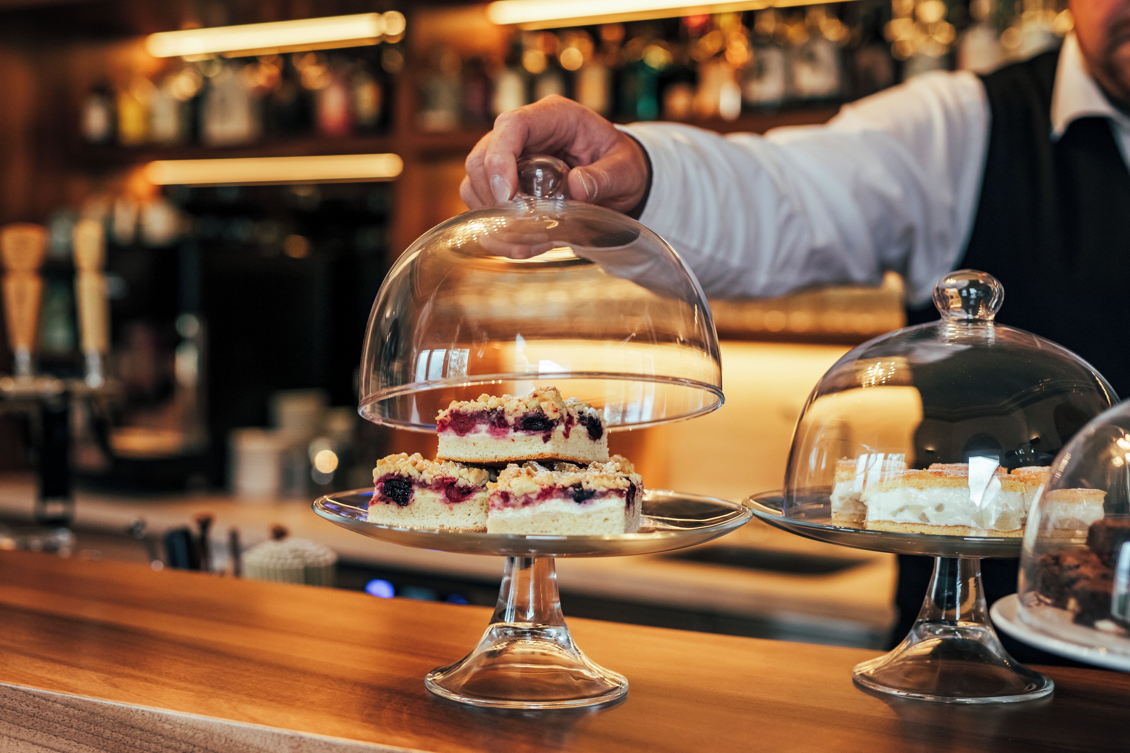 Una presentación de postres cubierta por una cúpula de cristal, una persona alcanzándola y un fondo desenfocado que sugiere un bar o restaurante.