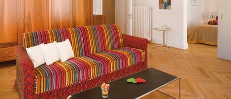 Un sofá colorido a rayas con cojines blancos, una mesa de centro negra con dos objetos rojos y verdes, y un suelo de madera.