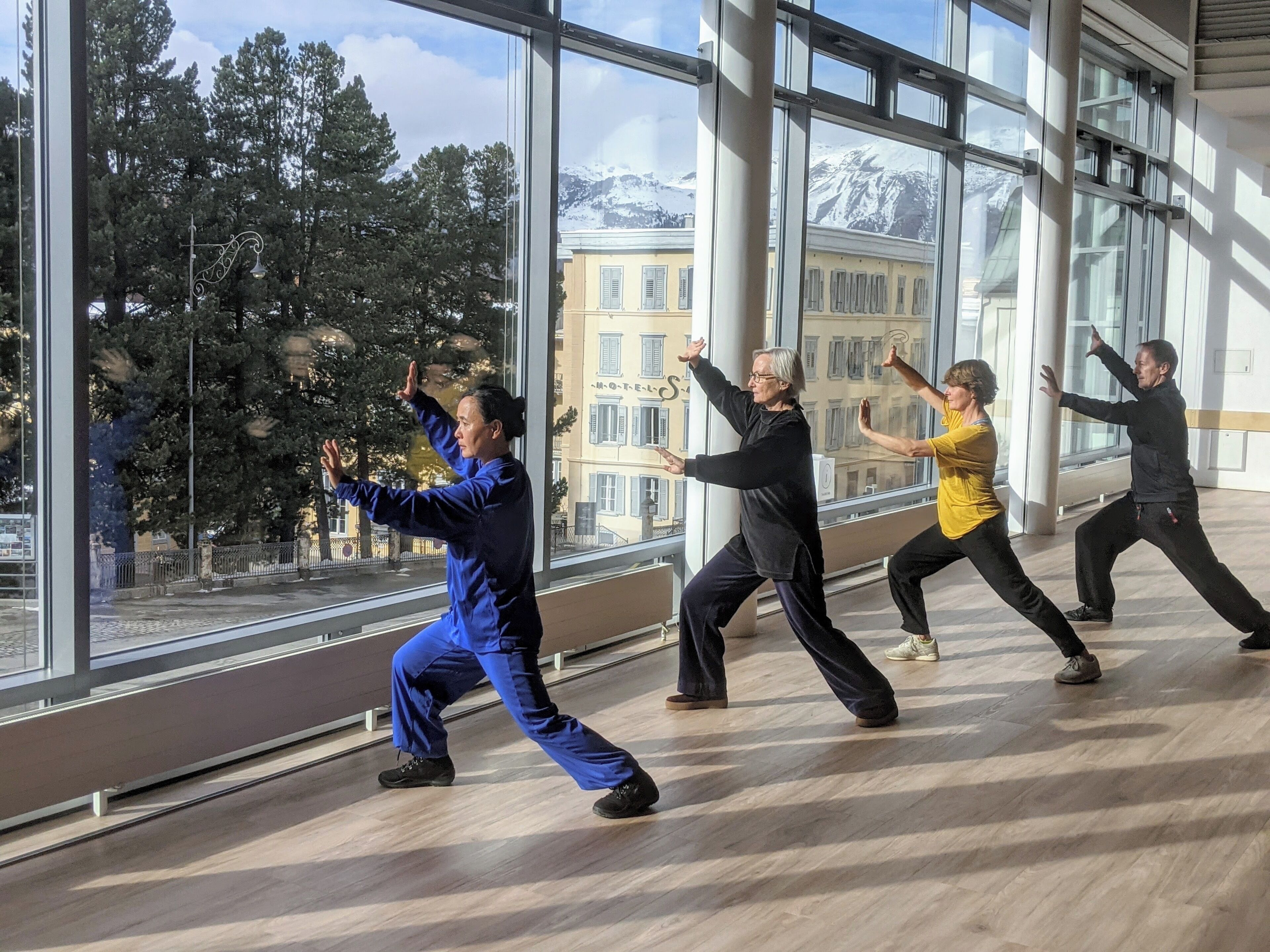 A group of people practicing Tai Chi in a spacious room with large windows offering a view of buildings and trees.