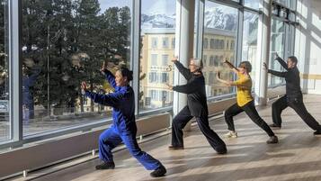 A group of people practicing Tai Chi in a spacious room with large windows offering a view of buildings and trees.