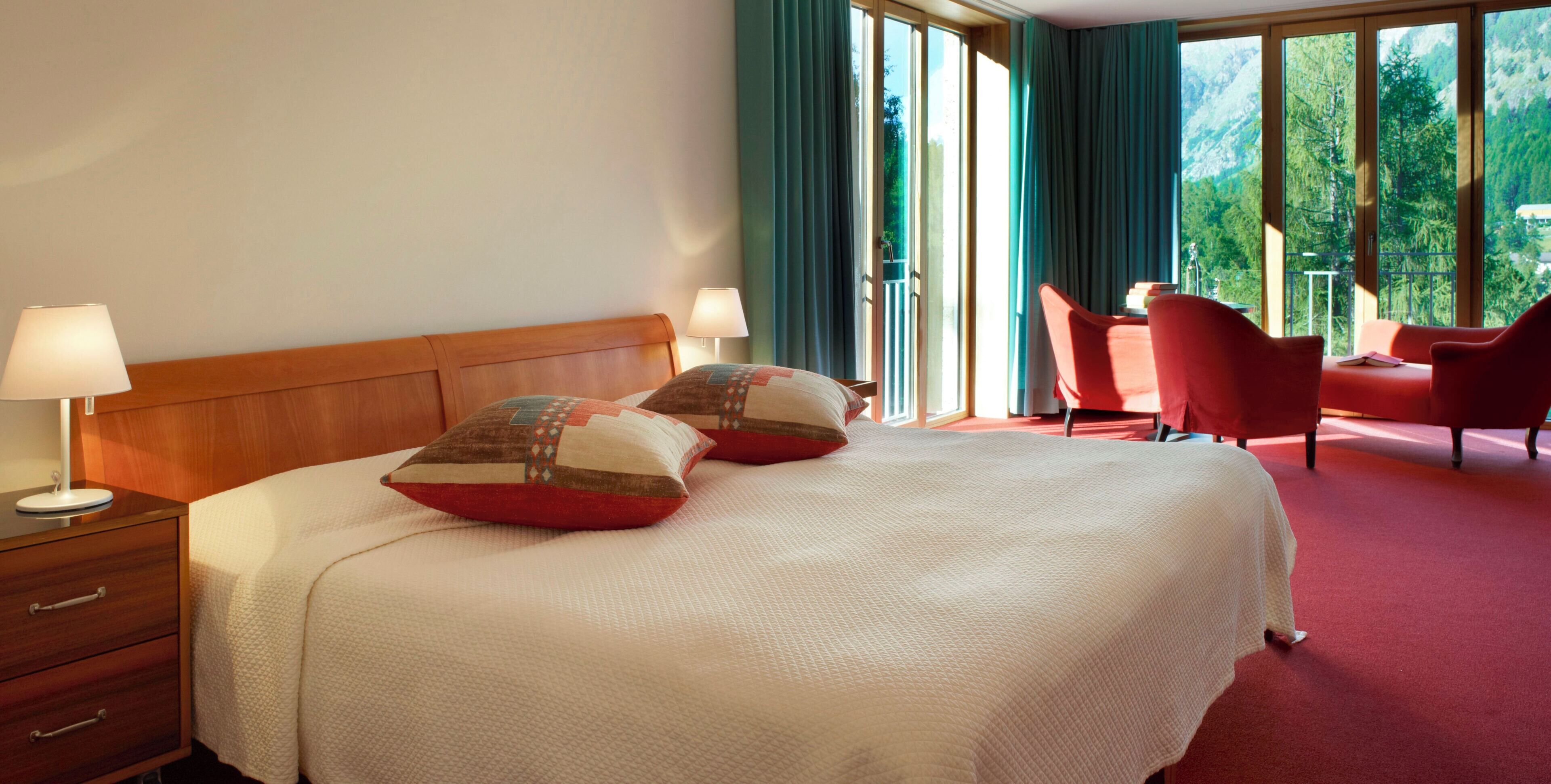Habitación de hotel con una cama grande, dos lámparas de noche, cabecera de madera, una silla roja y un balcón con vistas a árboles.