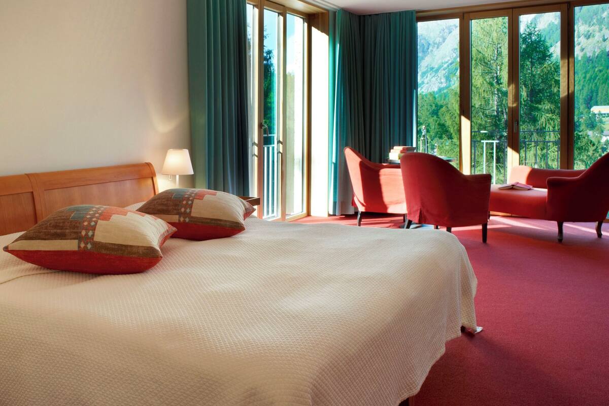 A hotel room with a large bed, two bedside lamps, a wooden headboard, a red chair, and a balcony with a view of trees.