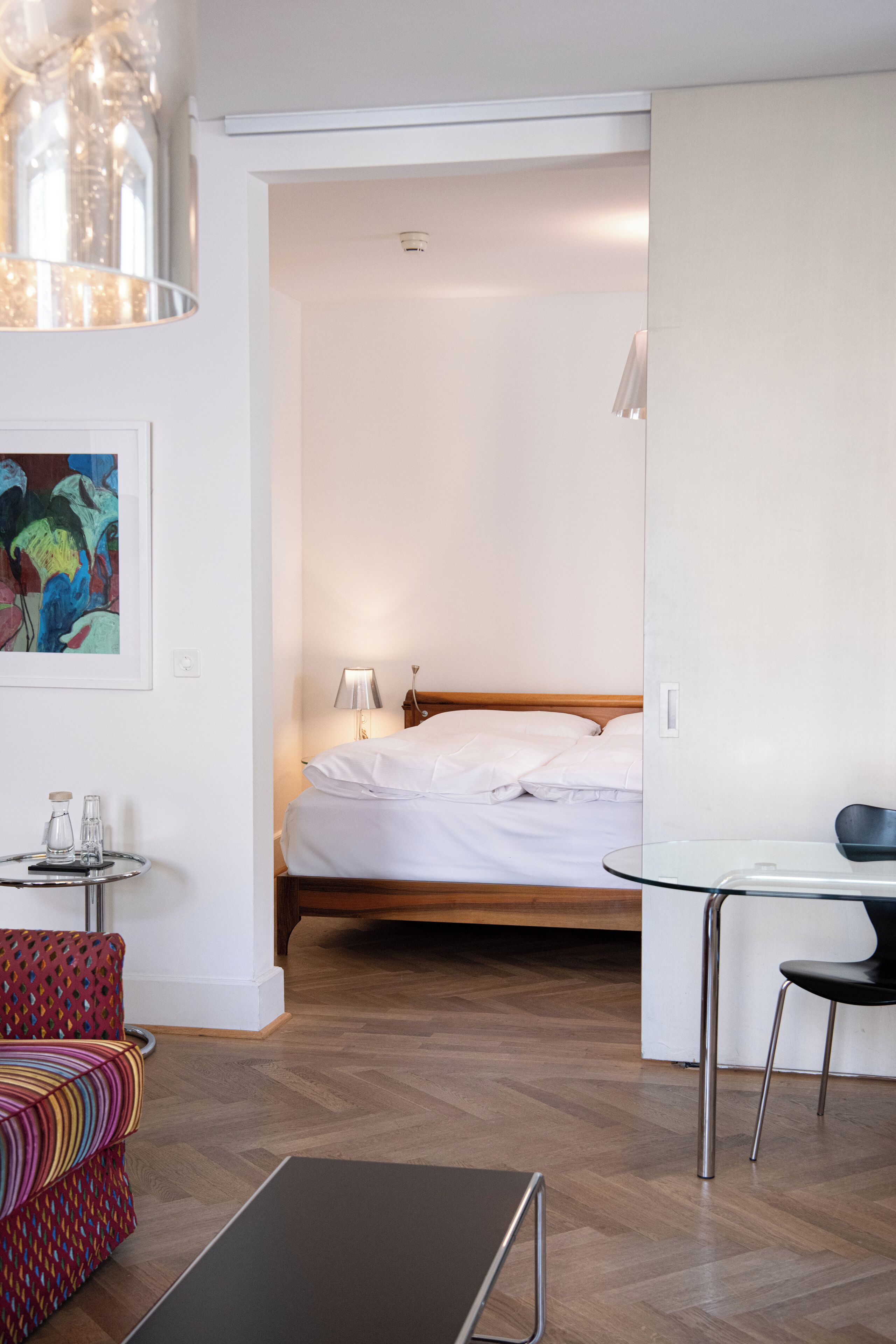 A modern hotel room with a wooden floor, a bed with white linens, a small round table with a chair, a colorful sofa, and a painting on the wall.