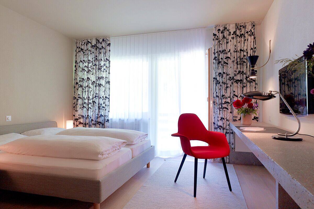 A bedroom with a bed, a desk with a chair, a lamp, and a window with curtains.