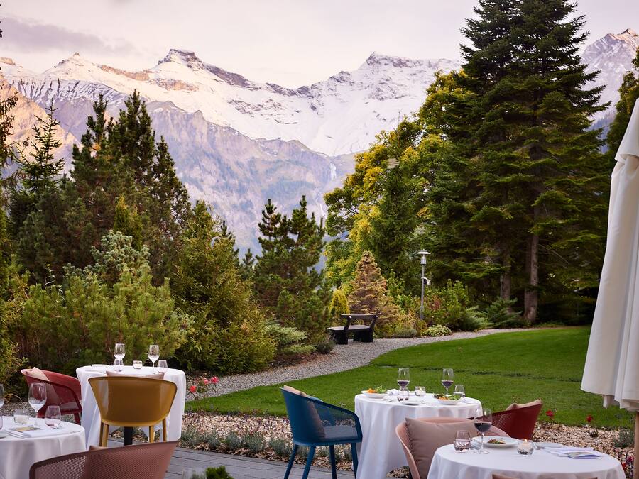 Restaurante con terraza al aire libre y vistas a montañas nevadas, rodeado de vegetación.