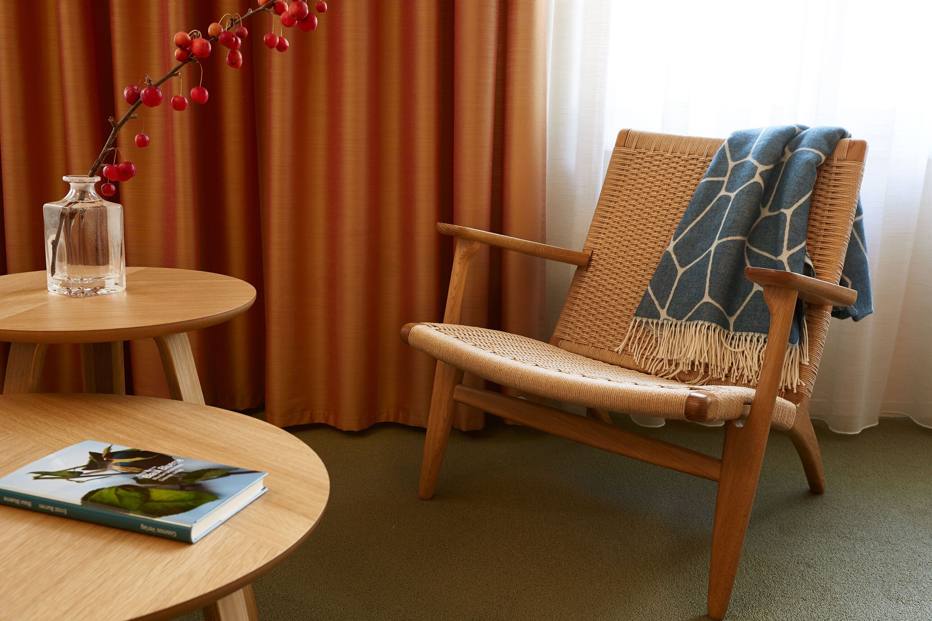 A wicker chair with a blue and white patterned throw blanket, a clear glass vase with red berries, and a book on a round wooden table.