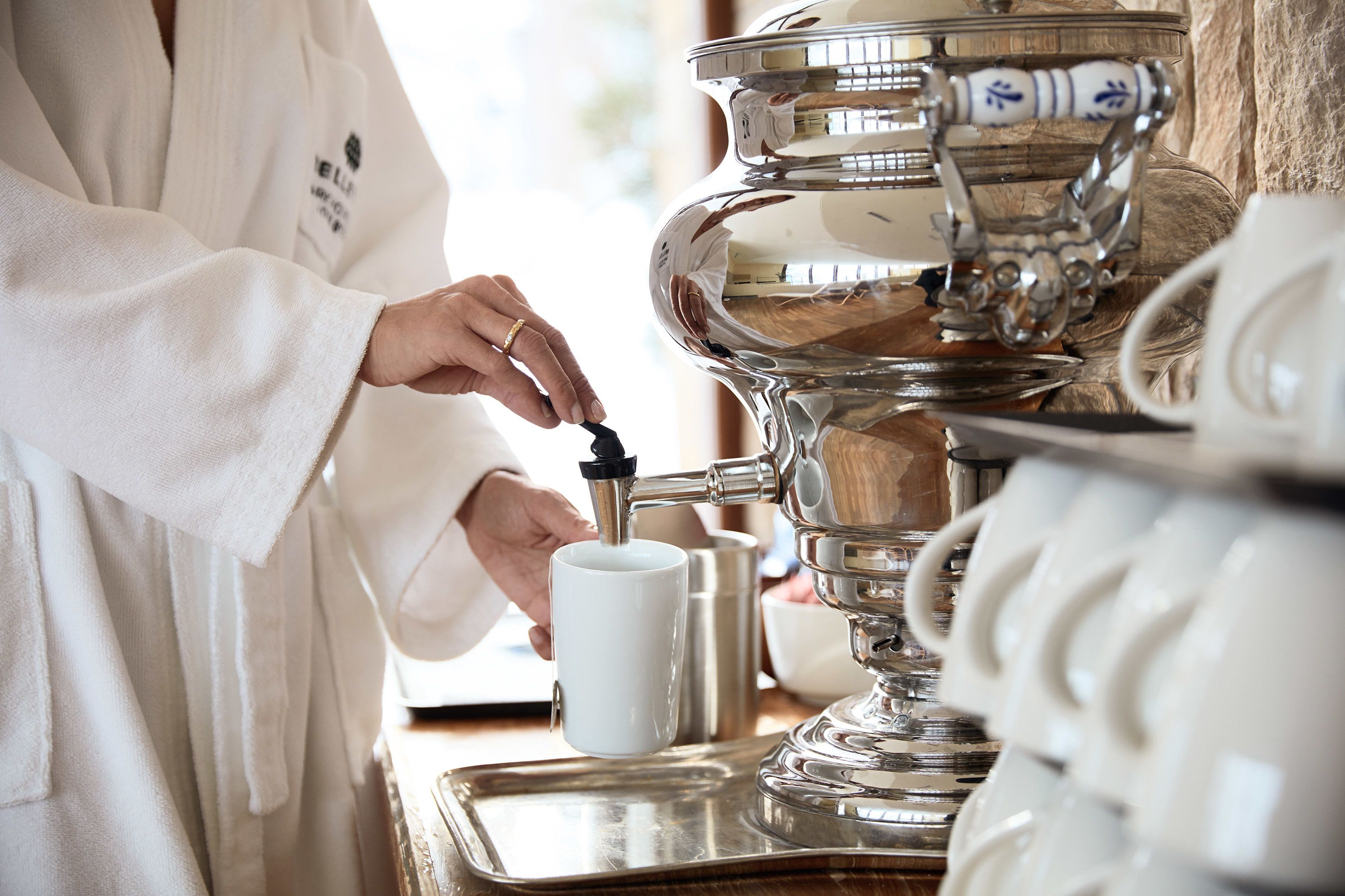 Une personne vêtue d’une robe blanche utilise une machine à café. On peut voir un plateau et une tasse à proximité.