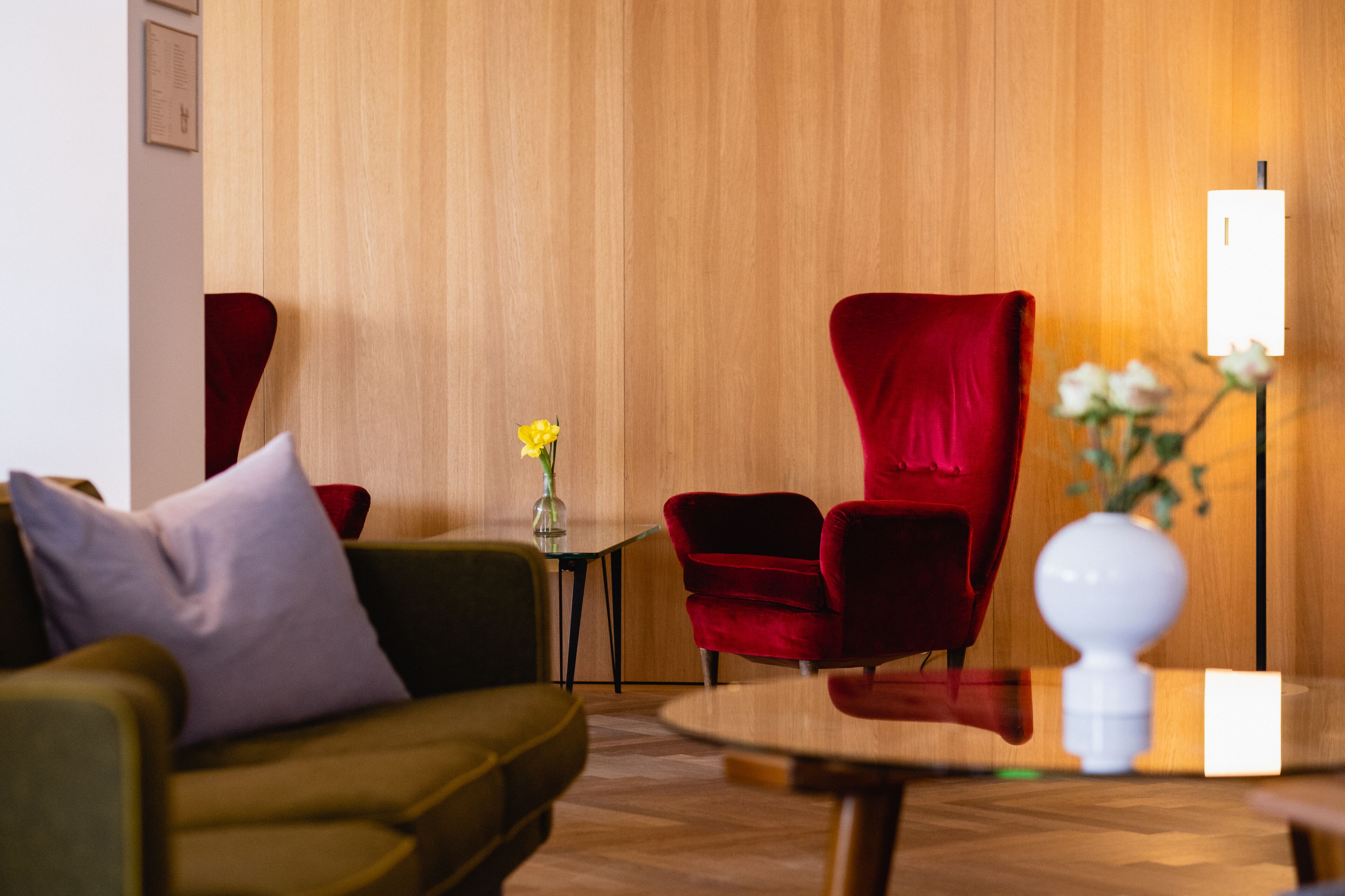 A modern hotel lobby with a wooden wall, a green sofa, a red armchair, a small table with a vase of flowers, and a floor lamp.