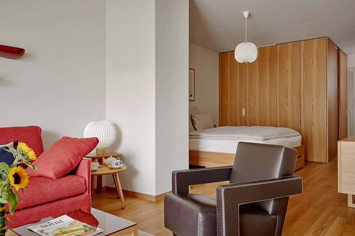A modern hotel room with a wooden wardrobe, a bed, a red sofa, a brown leather chair, and a small round table with a lamp and teapot.