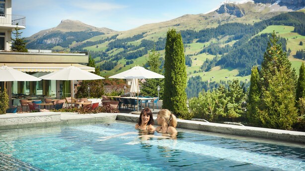 A swimming pool with two people, surrounded by outdoor seating and umbrellas, overlooking a mountainous landscape. - Bellevue Parkhotel & Spa - Relais & Châteaux (Adelboden)