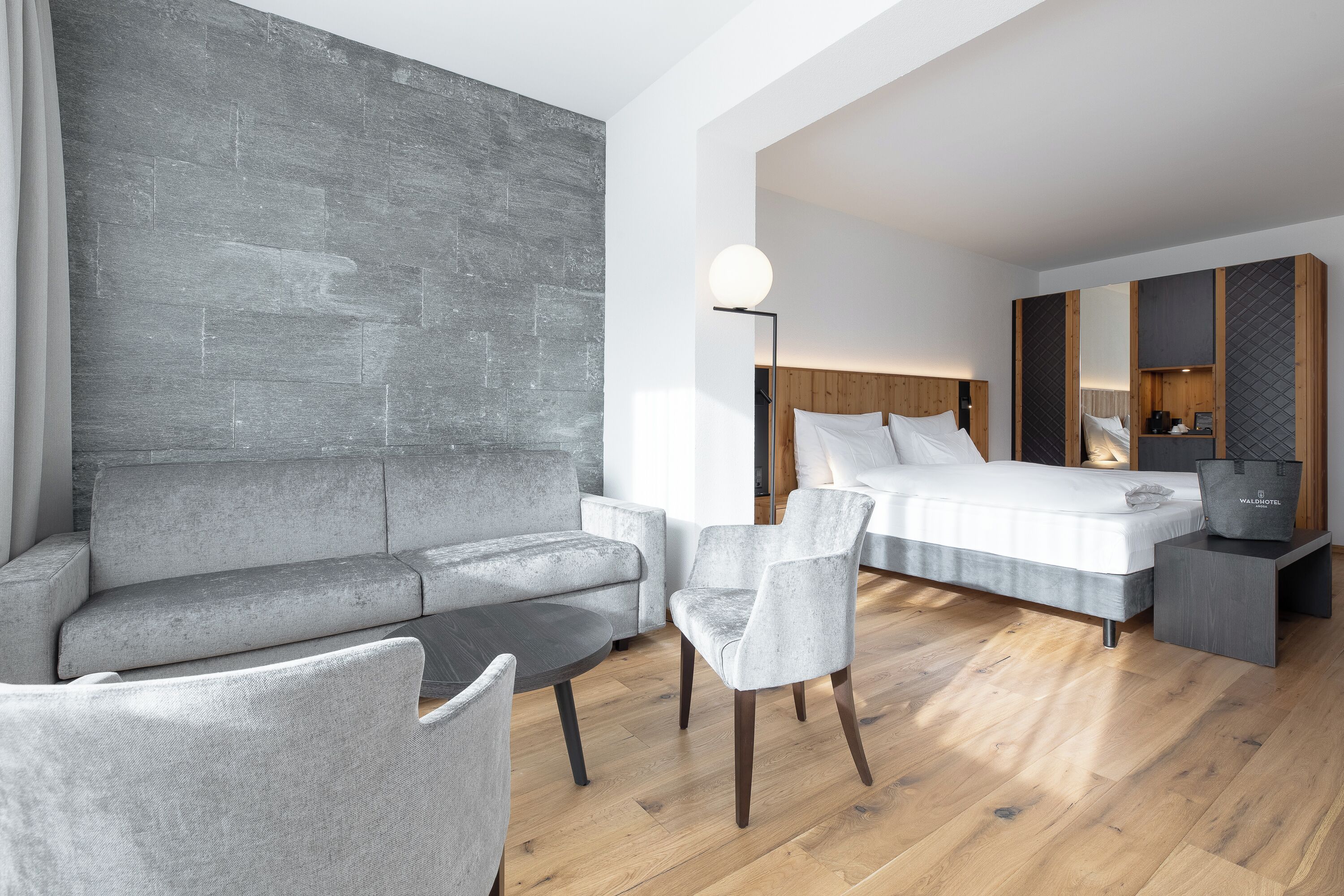 Une chambre d’hôtel moderne avec un canapé gris, une table basse ronde, un fauteuil gris, un plancher en bois, un lit avec une literie blanche, une table de chevet, une lampe et un miroir.