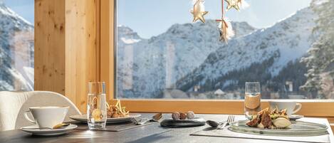 Une table à manger, dressée avec des assiettes, des couverts et des verres, placée devant une fenêtre offrant une vue sur les montagnes.