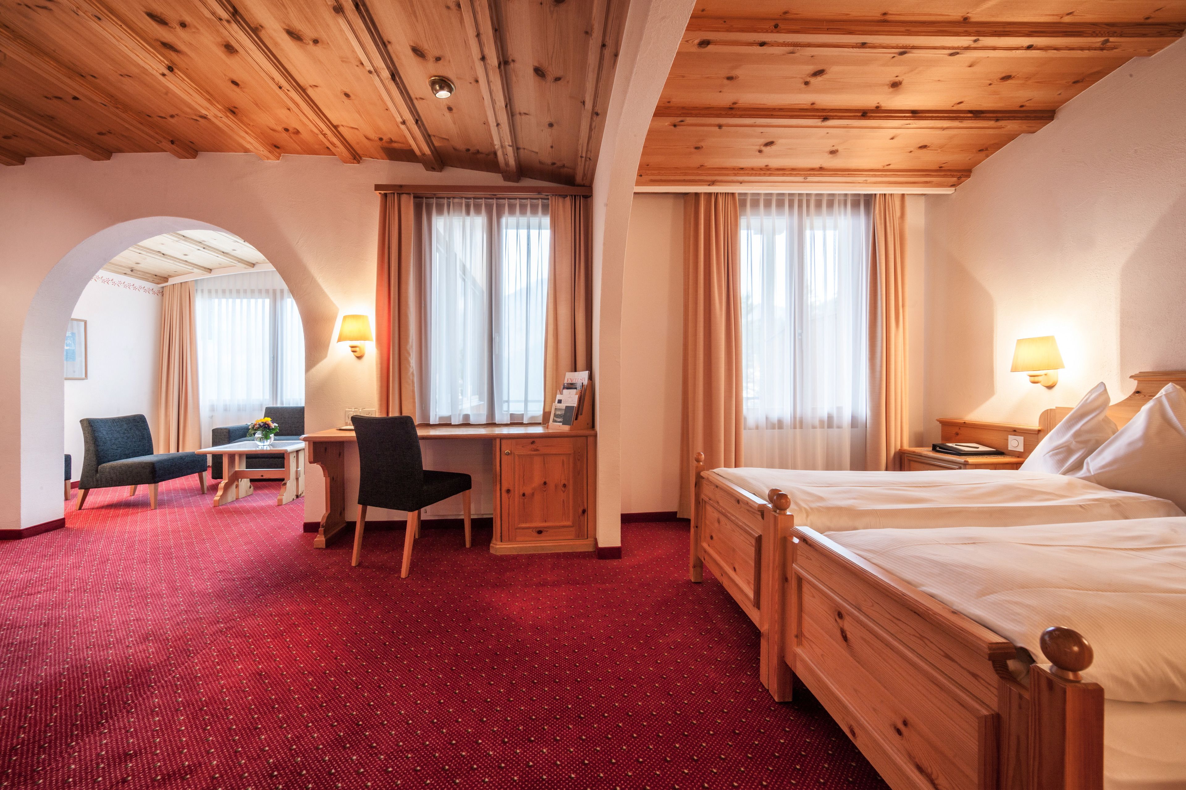 A hotel room with a wooden ceiling, two beds, a desk with a chair, and a red carpeted floor.