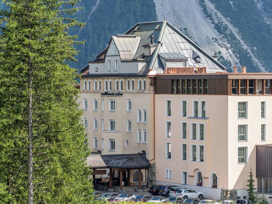 A multi-story hotel with a mountain backdrop, featuring a parking area and a covered outdoor seating area.