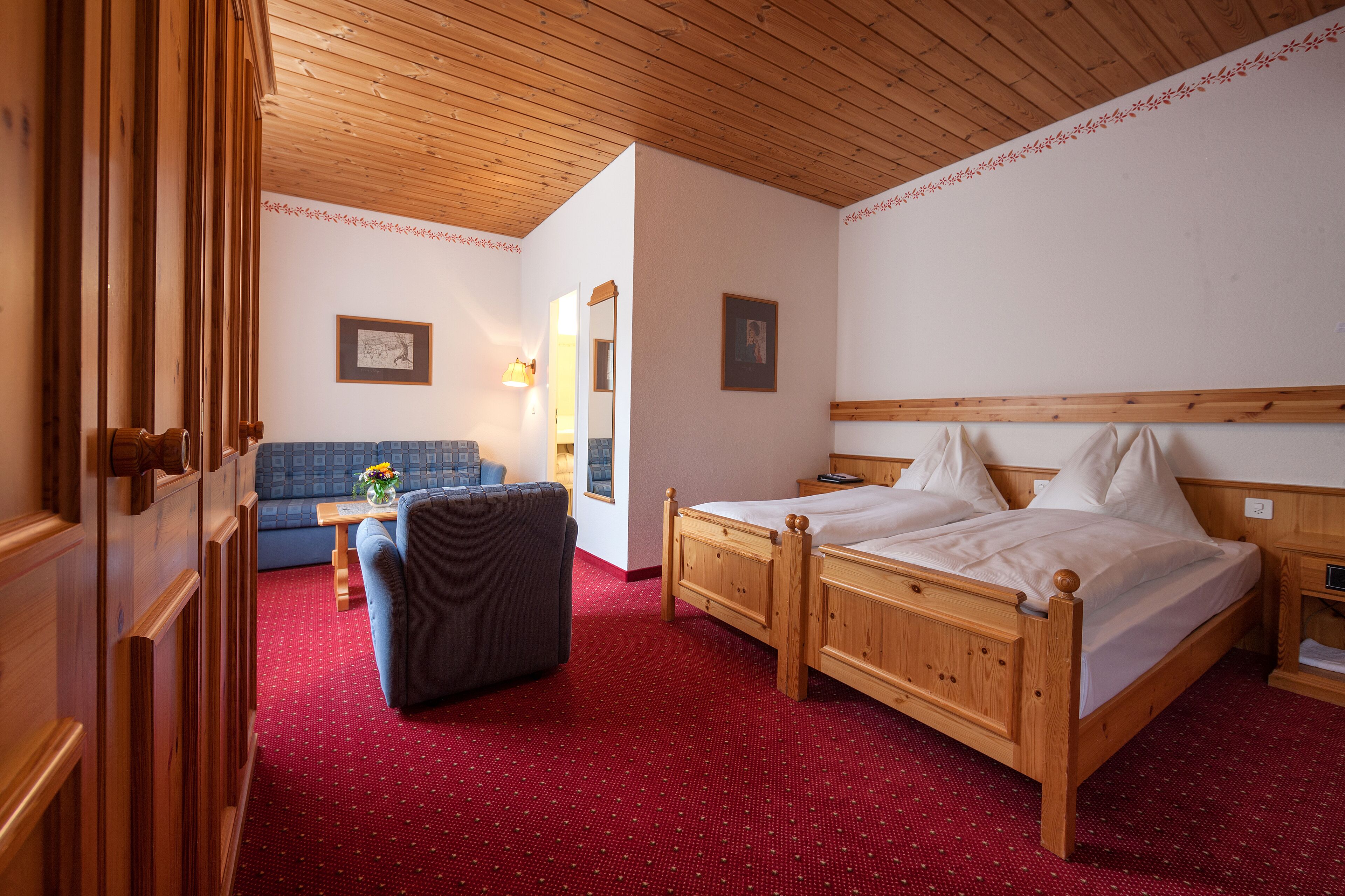 A wooden-framed bed with white linens, a dark armchair, and a red carpeted floor in a room with wooden ceiling beams and white walls.