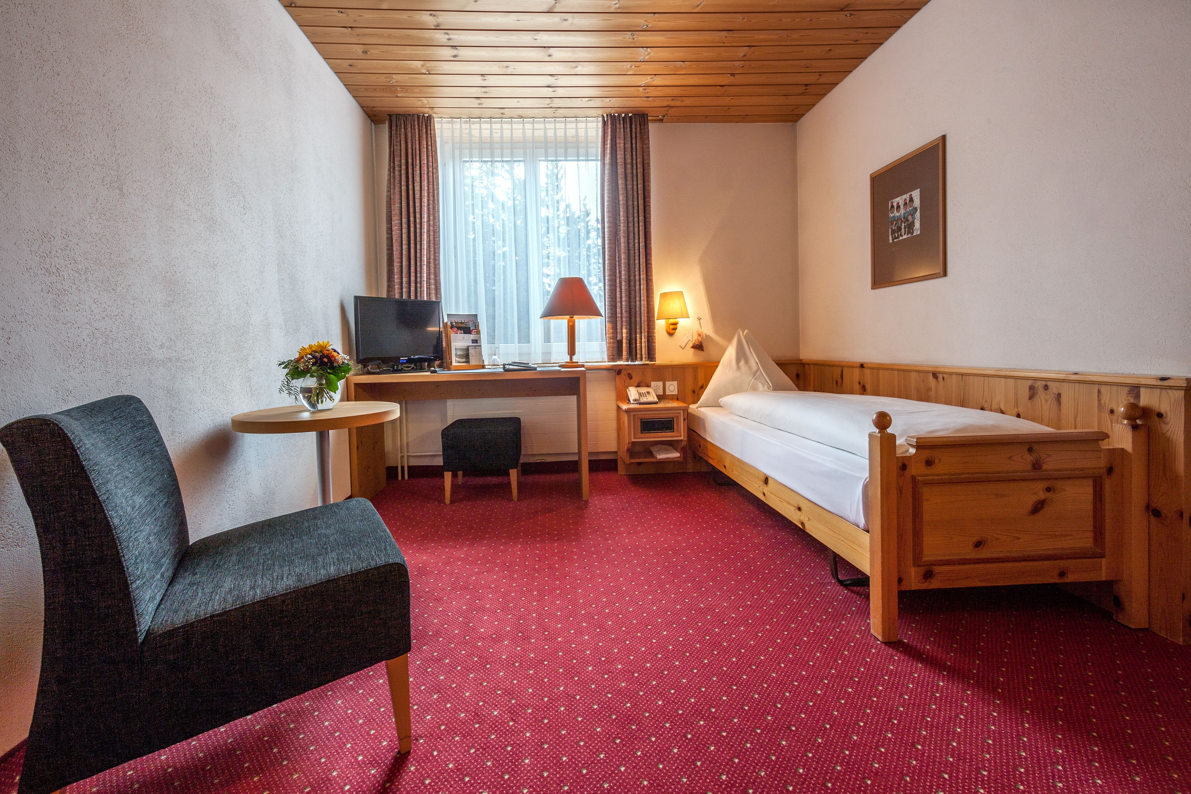 Une chambre d’hôtel avec un lit en bois, un bureau avec un ordinateur, une chaise, une petite table et une lampe.