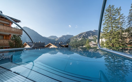 A clear view of a swimming pool with a mountain backdrop, wooden buildings, and a glass balustrade.