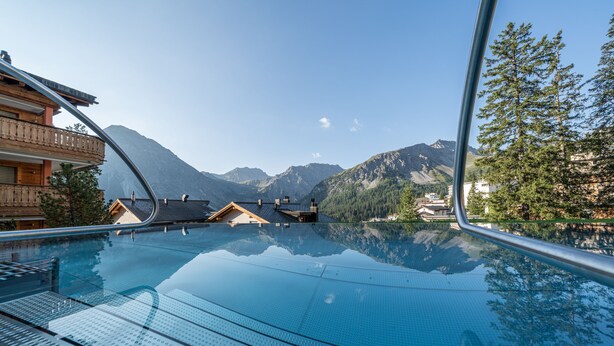 Vista despejada de una piscina con montañas al fondo, edificios de madera y barandilla de vidrio.