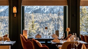 Un comedor con mesas dispuestas para dos, grandes ventanas que ofrecen vistas a un bosque nevado y montañas.