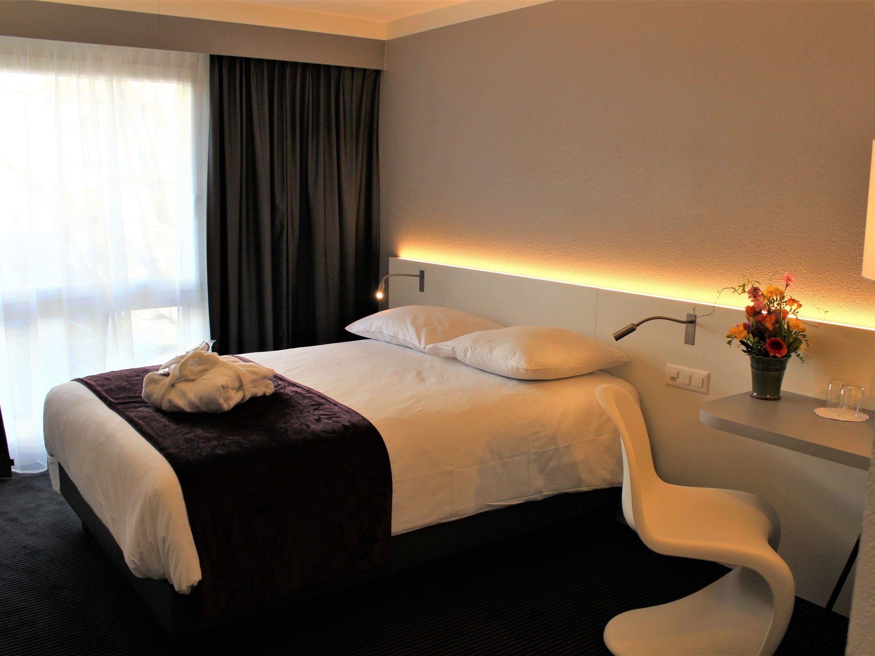 Habitación de hotel con cama, mesita de noche con un florero, una silla y una ventana con cortinas.