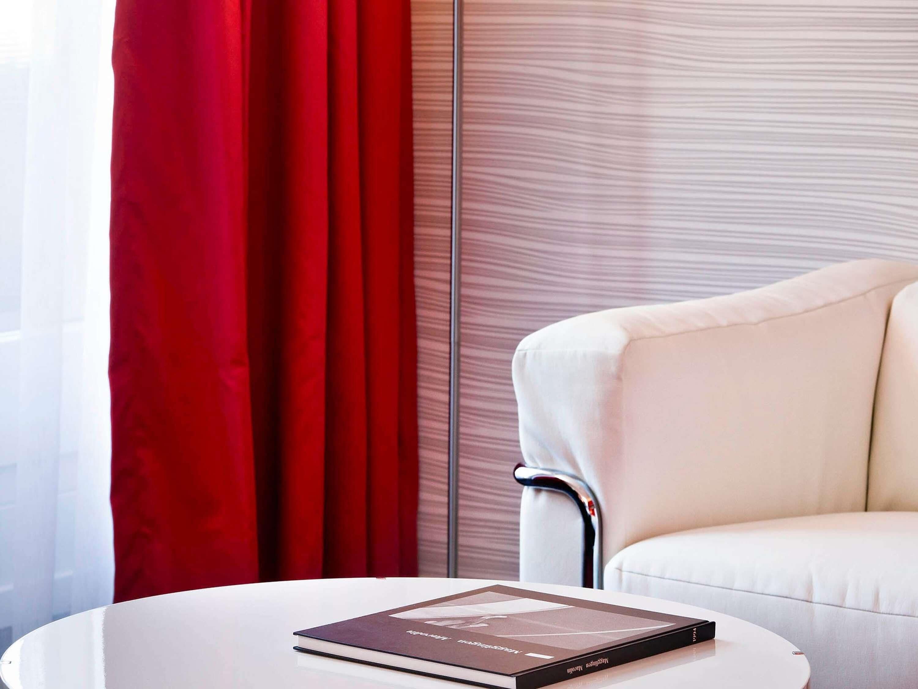 Ein modernes Zimmer mit einem weißen Ledersessel, einem runden weißen Tisch mit einem Buch, roten Vorhängen und einer gestreiften Wand.