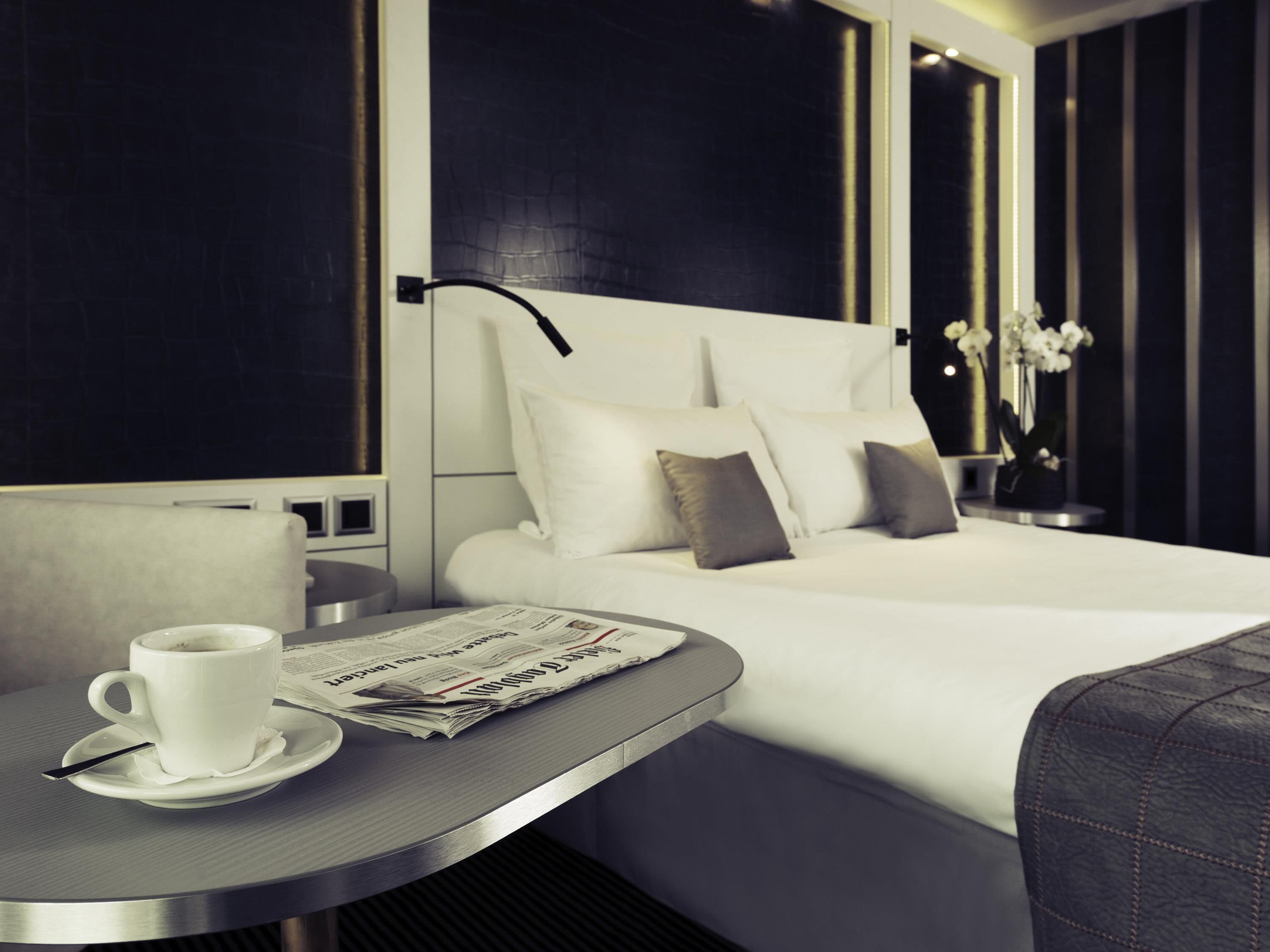 Ein modernes Hotelzimmer mit einem ordentlich bezogenem Bett, einem Nachttisch mit Tasse und Zeitung sowie einer Vase mit Blumen.