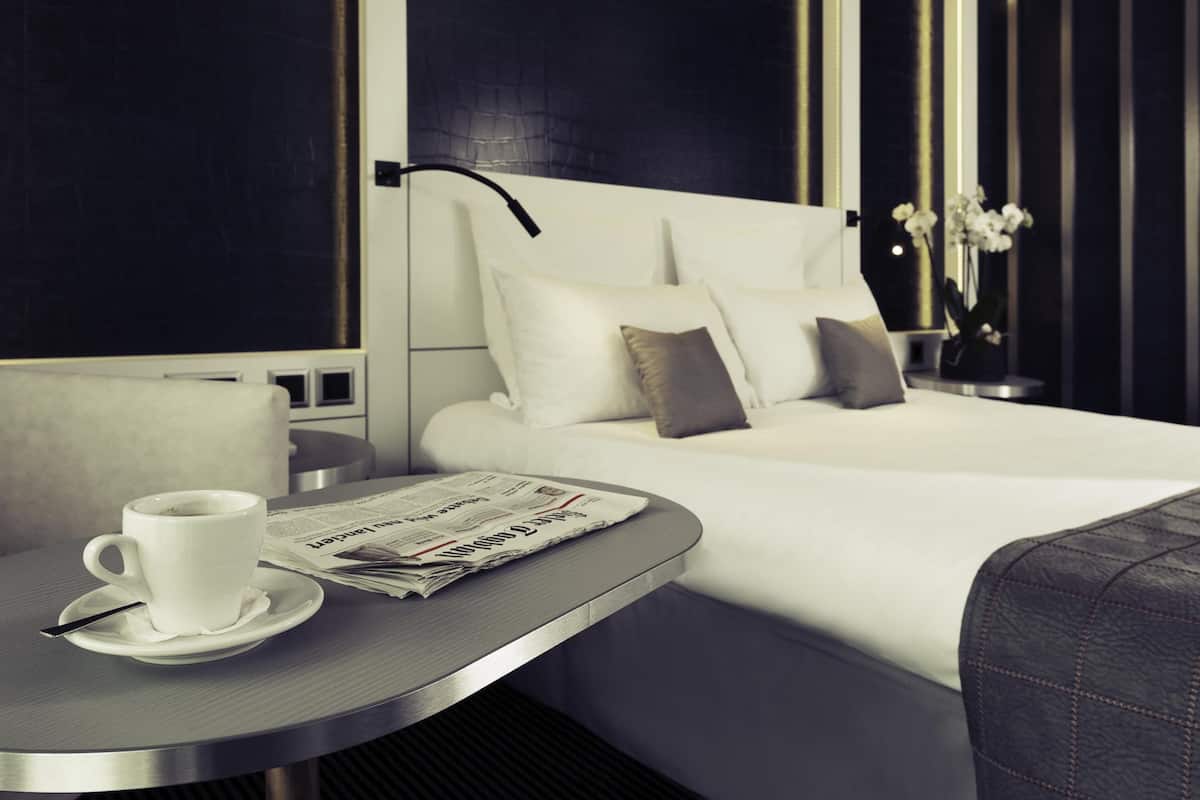 Ein modernes Hotelzimmer mit einem ordentlich bezogenem Bett, einem Nachttisch mit Tasse und Zeitung sowie einer Vase mit Blumen.
