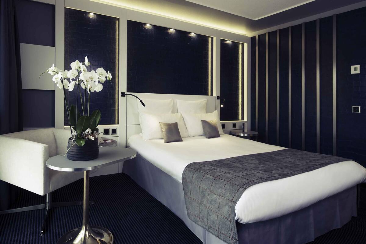 A modern hotel room with a large bed, a bedside table, a chair, and a potted plant.