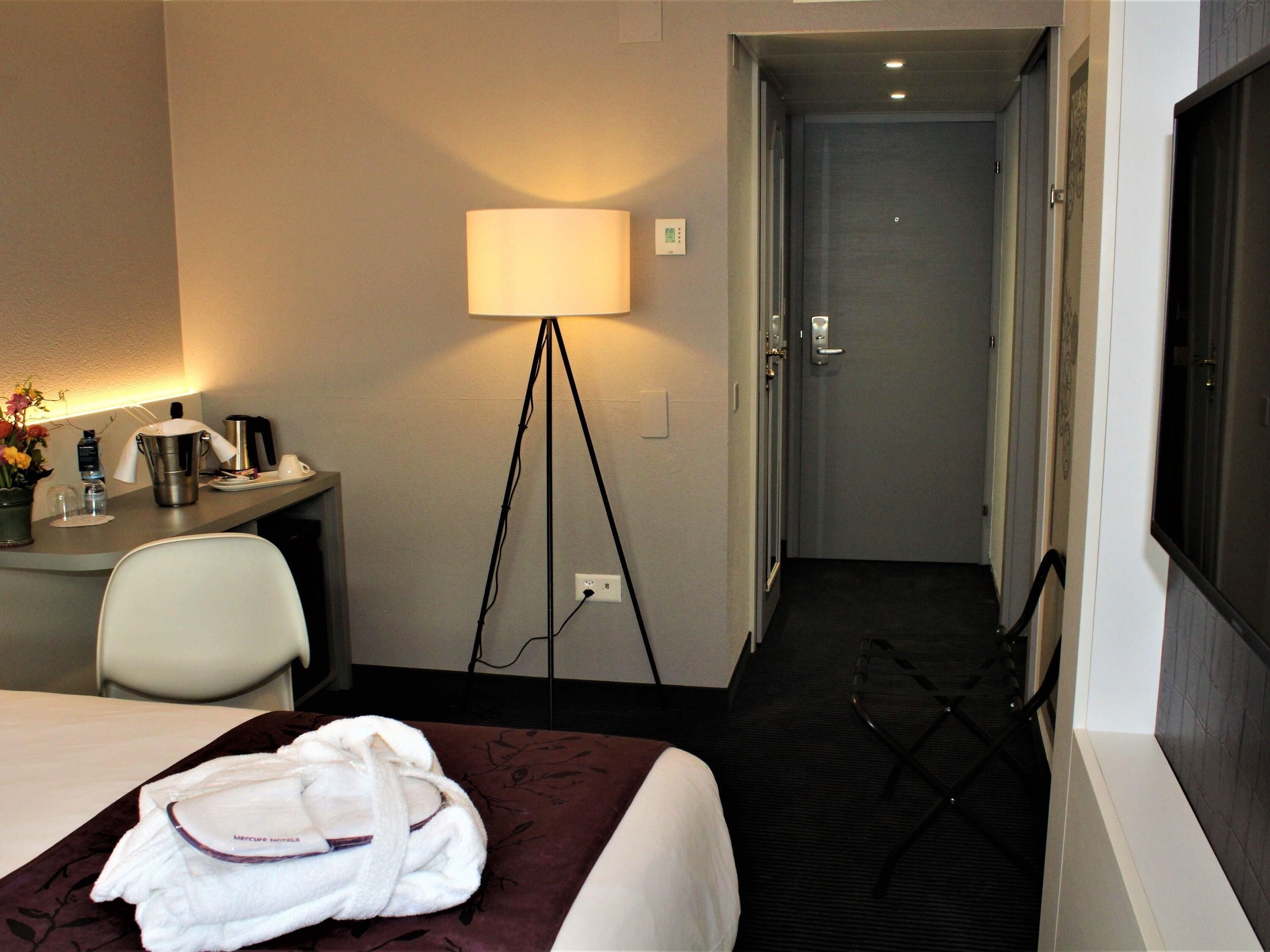 A hotel room with a bed, a desk with a lamp, a chair, a robe, and a bathroom visible through an open door.