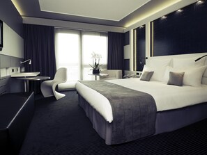 Une chambre d’hôtel moderne avec un grand lit, un bureau avec une chaise, une petite table avec une plante et une grande fenêtre avec des rideaux.