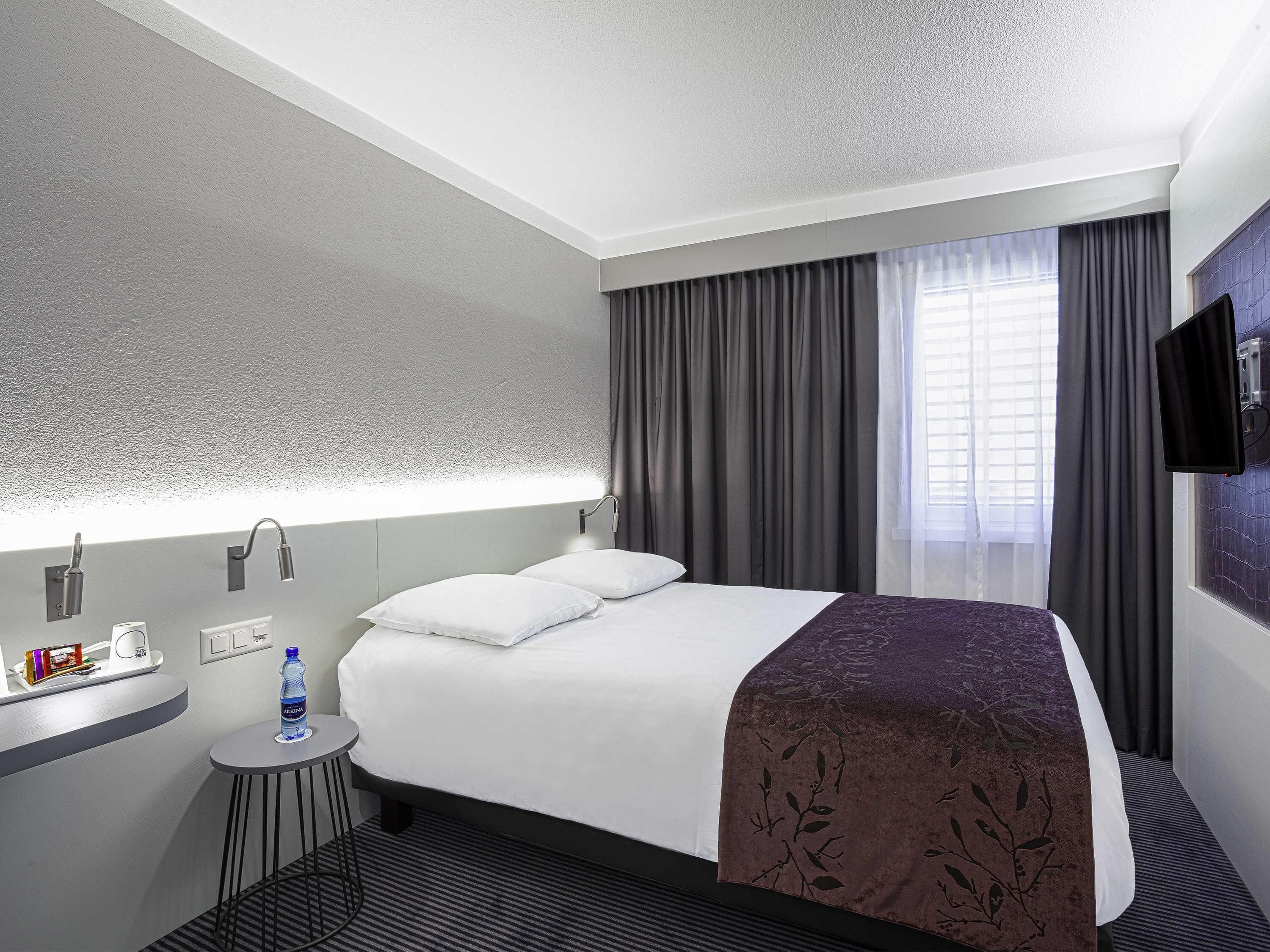 A hotel room with a bed, a small table, a water bottle, a TV, and a window with curtains.