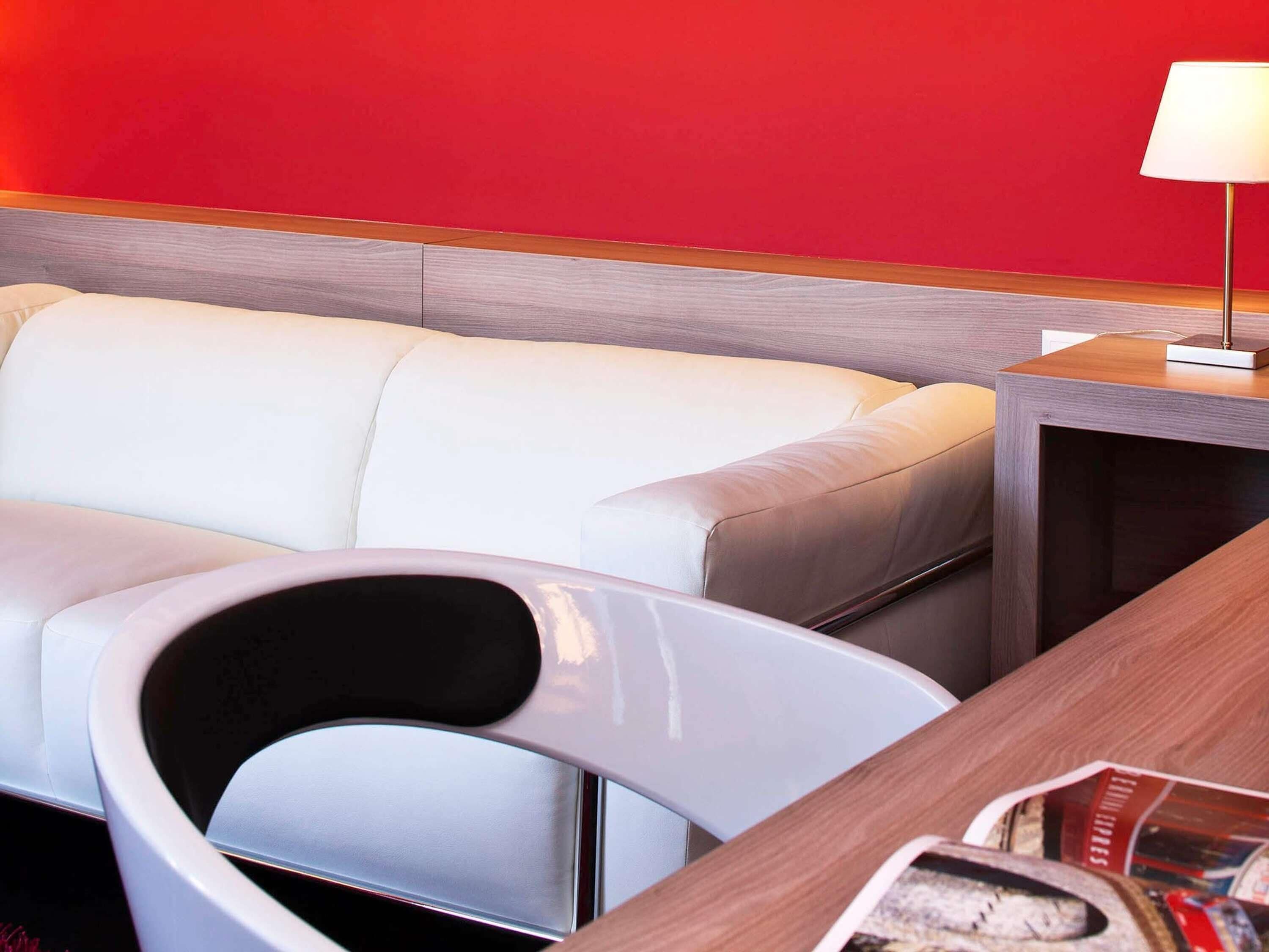 A modern hotel room with a white leather sofa, a wooden side table with a lamp, and a magazine on the table.