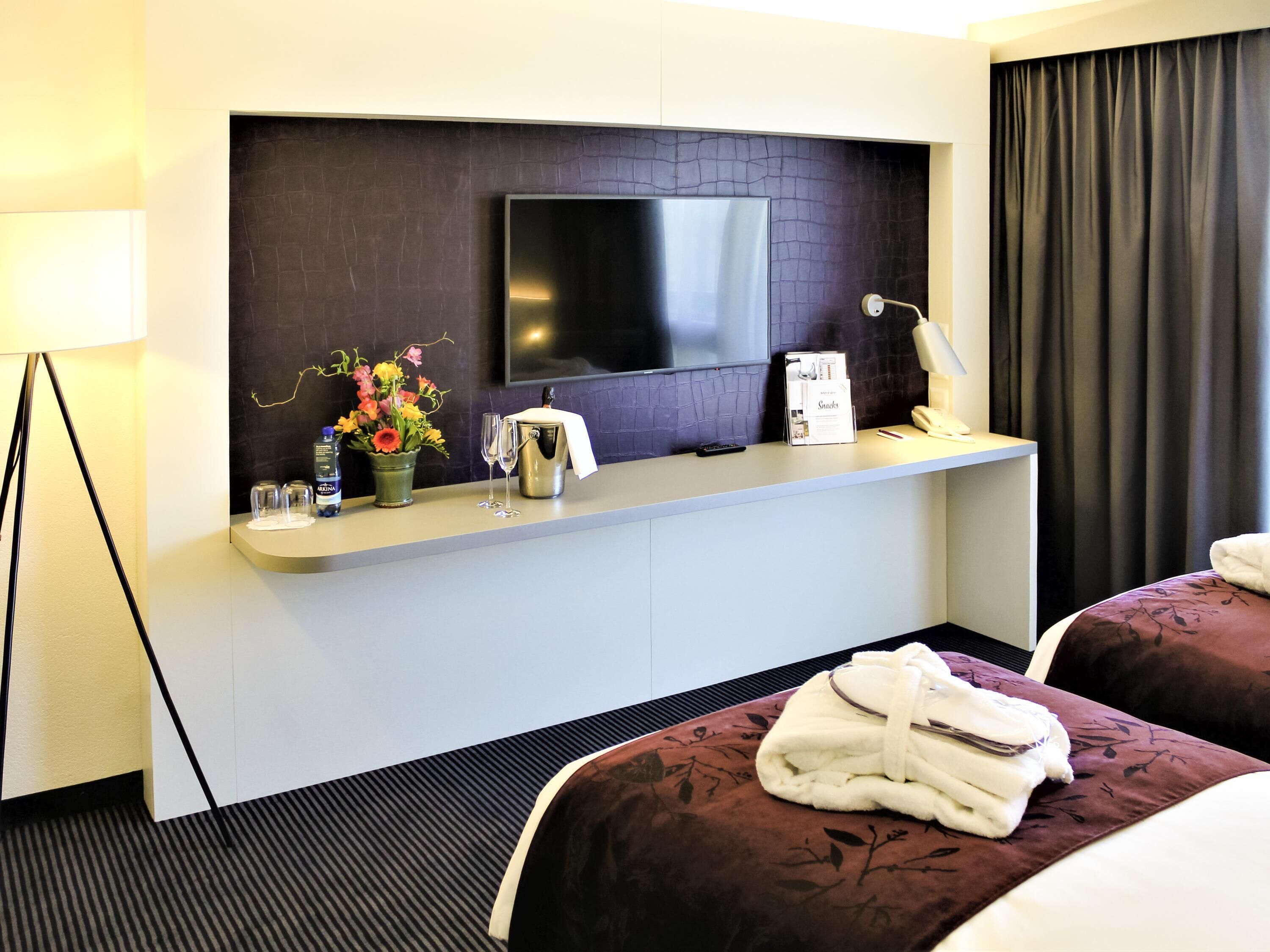 Ein Hotelzimmer mit einem Flachbildfernseher an einer dunkel strukturierten Wand, einem kleinen Tisch mit Blumen, einer Wasserflasche und einer Minibar. Es gibt zwei Betten mit dunkler Bettwäsche und jeweils ein Paar Hausschuhe.