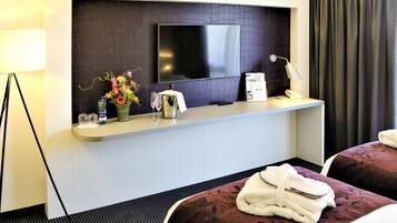 Ein Hotelzimmer mit einem Flachbildfernseher an einer dunkel strukturierten Wand, einem kleinen Tisch mit Blumen, einer Wasserflasche und einer Minibar. Es gibt zwei Betten mit dunkler Bettwäsche und jeweils ein Paar Hausschuhe.