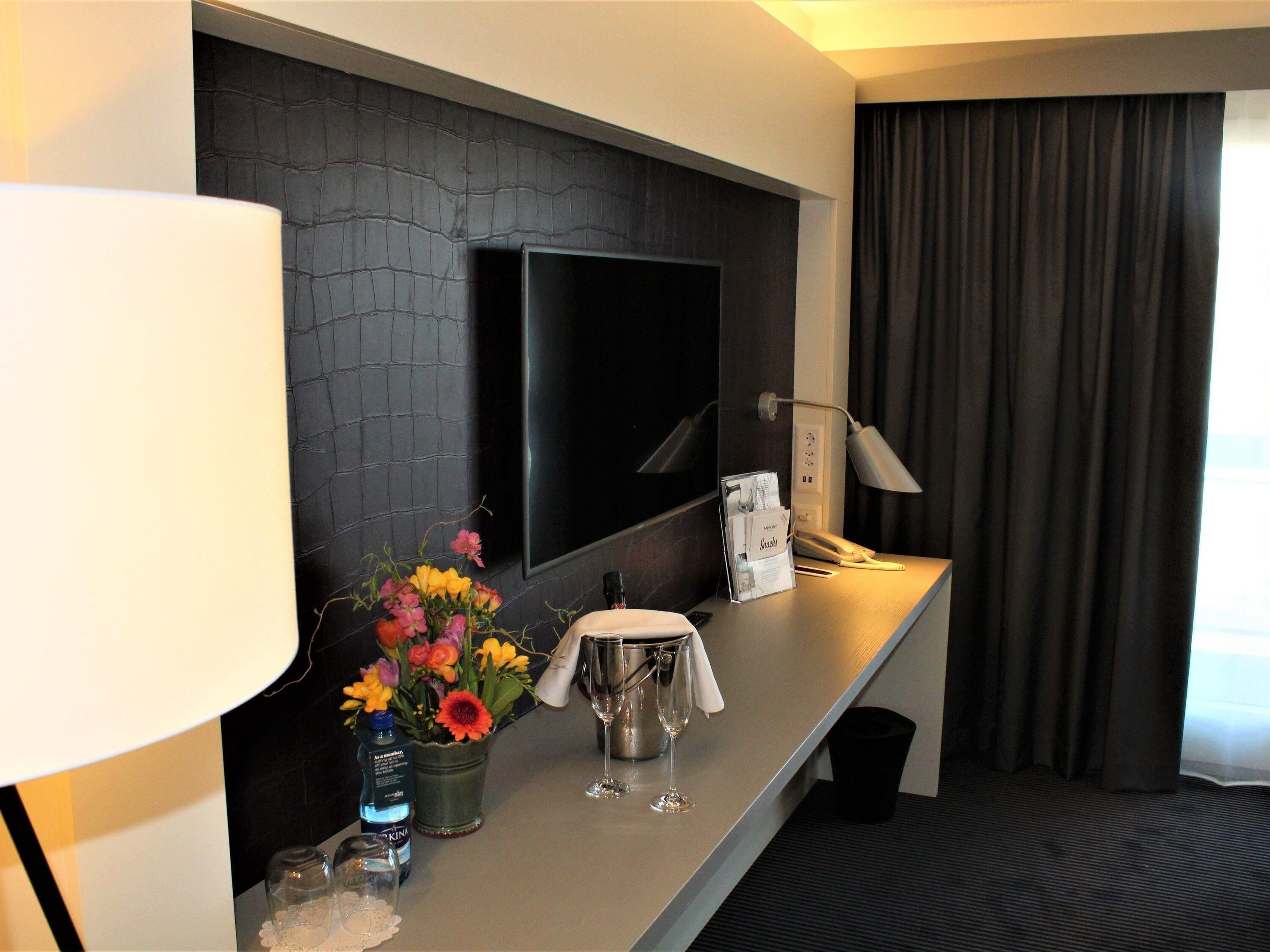 A hotel room with a flat-screen TV mounted on a textured wall, a desk with a lamp, a vase of flowers, a minibar, and a window with curtains.