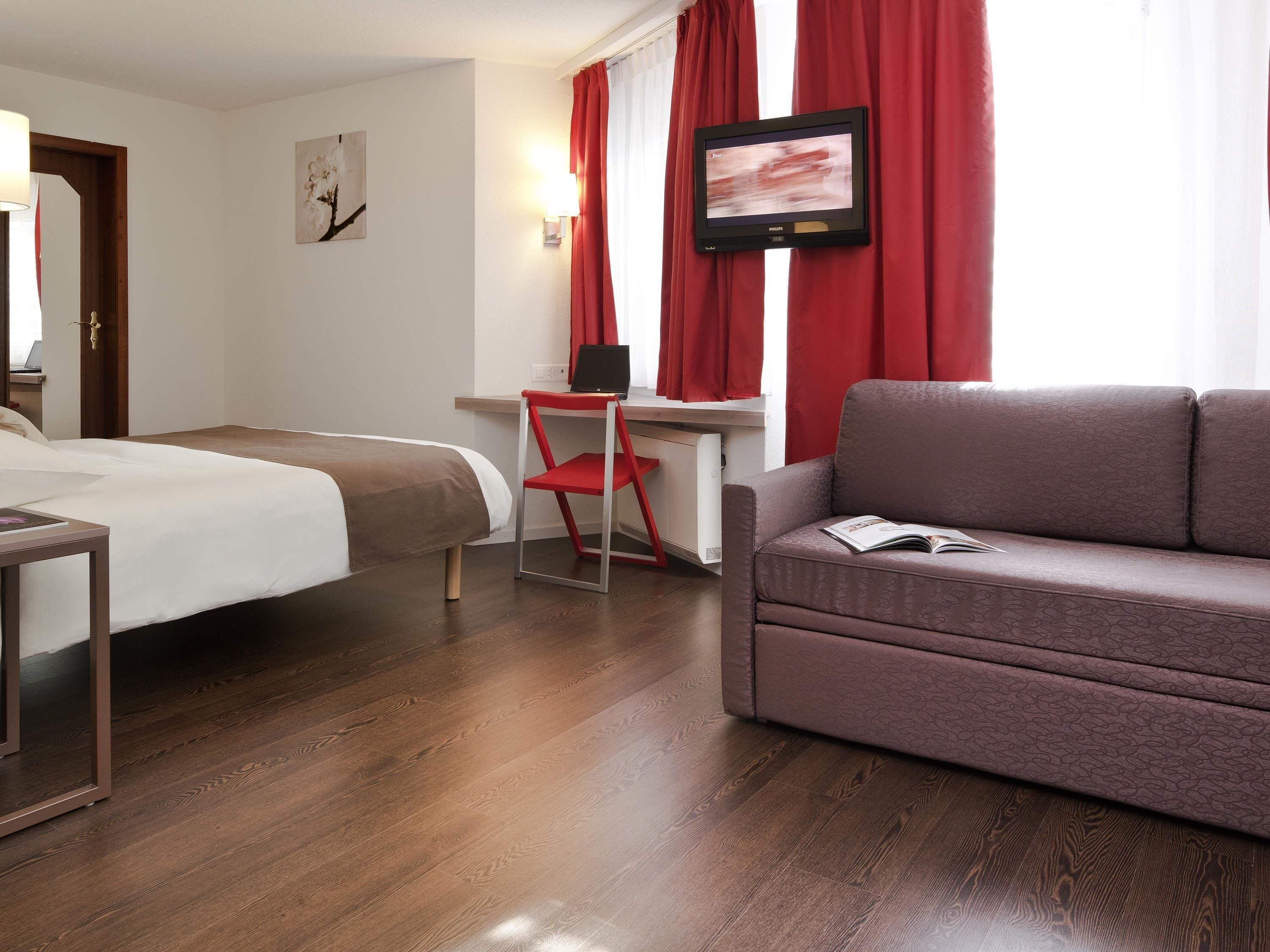 Ein Hotelzimmer mit einem Bett, einem Schreibtisch mit Stuhl, einer Couch und einem an der Wand befestigten Fernseher.