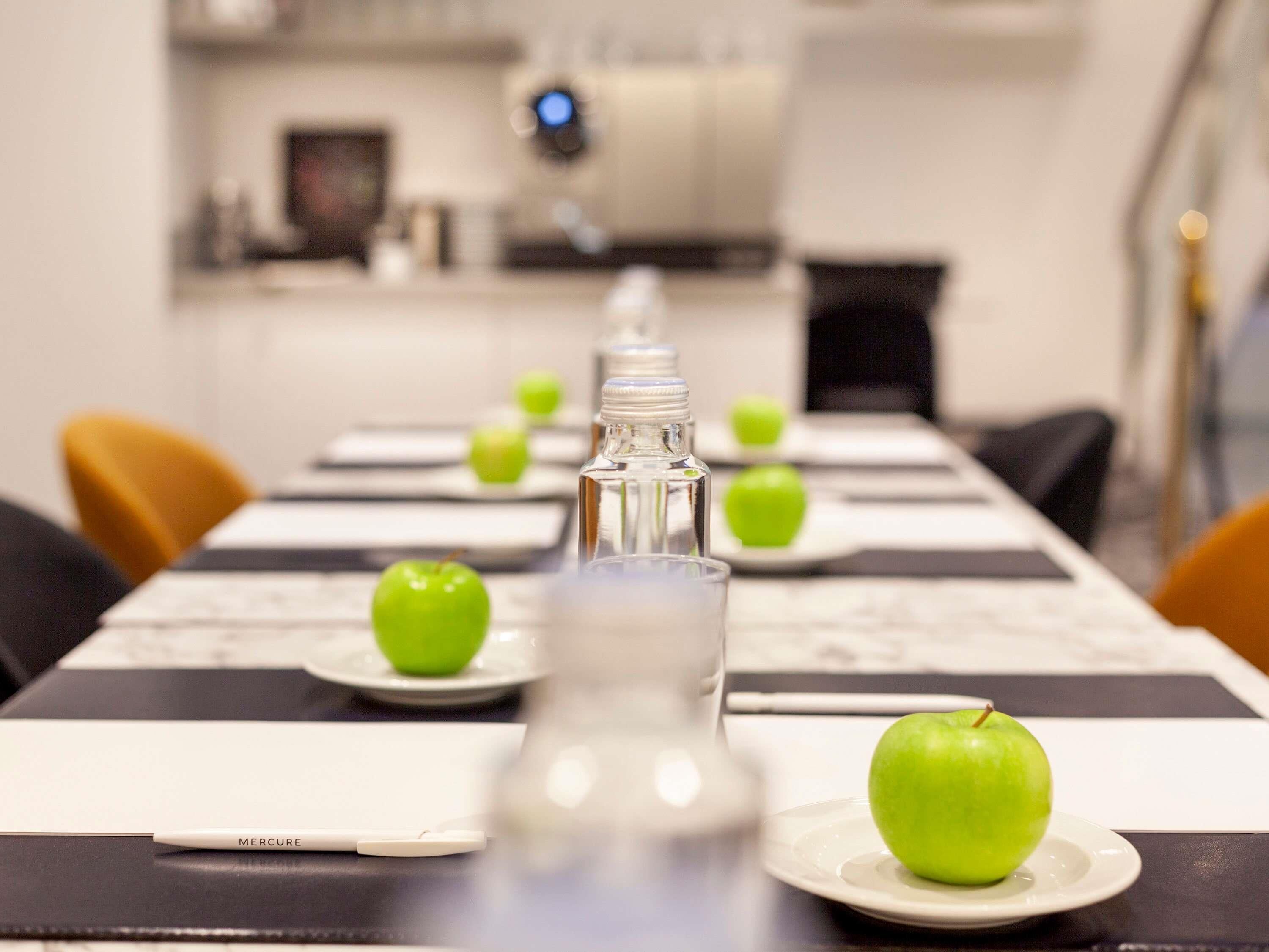 Una mesa de comedor con una botella, manzanas verdes y un mantel individual blanco con la etiqueta 'MERCURE'.