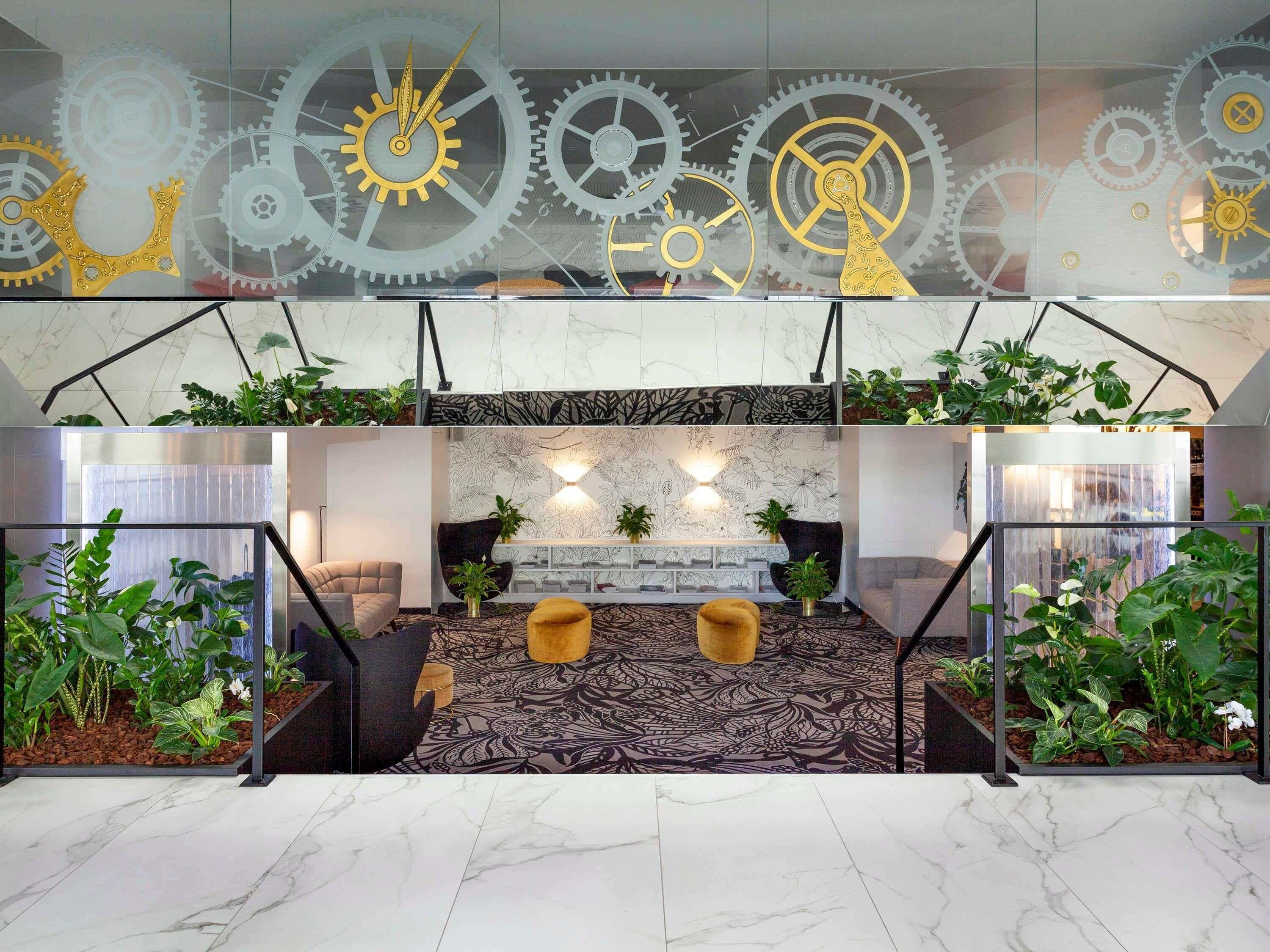 Eine Lobby im modernen Stil, mit Marmorboden, einer zentralen Sitzecke mit Sofa und Hocker sowie einer Wand mit dekorativen Zahnrädern.