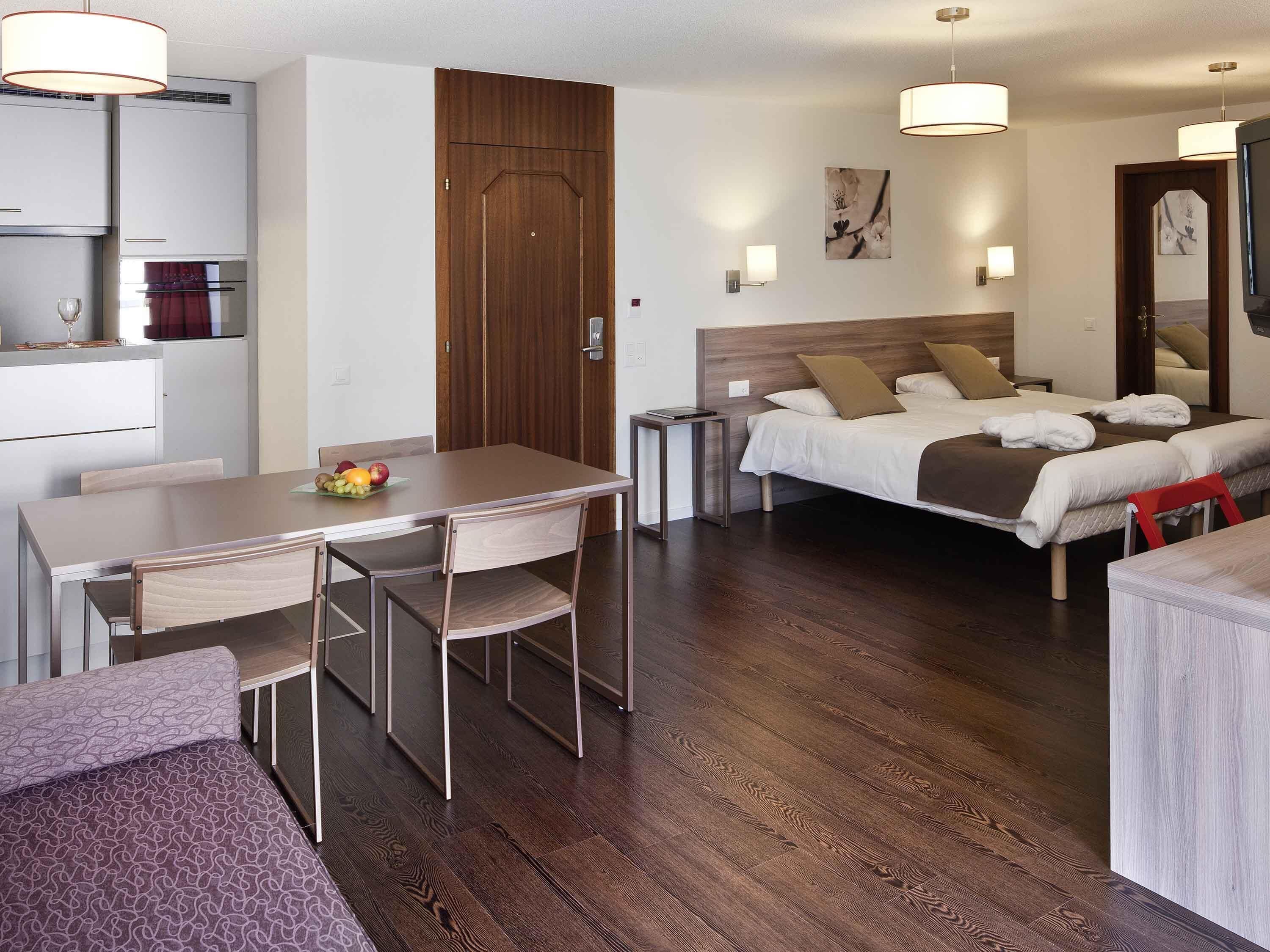 Une chambre d’hôtel comprenant un lit, des tables de chevet, un bureau avec des chaises, une petite kitchenette et un plancher en bois.