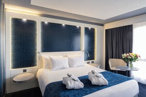 Una habitación de hotel moderna con una cama grande, dos mesitas de noche, una pequeña mesa redonda y una silla. Hay un jarrón con flores sobre la mesa.