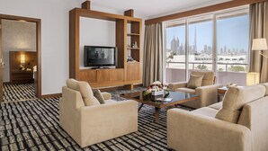 A hotel room with a sofa set, a television, a coffee table with flowers, and a city view through large windows.