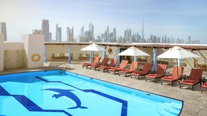 A rooftop pool with lounge chairs and umbrellas, a city skyline in the background, and a clear sky.