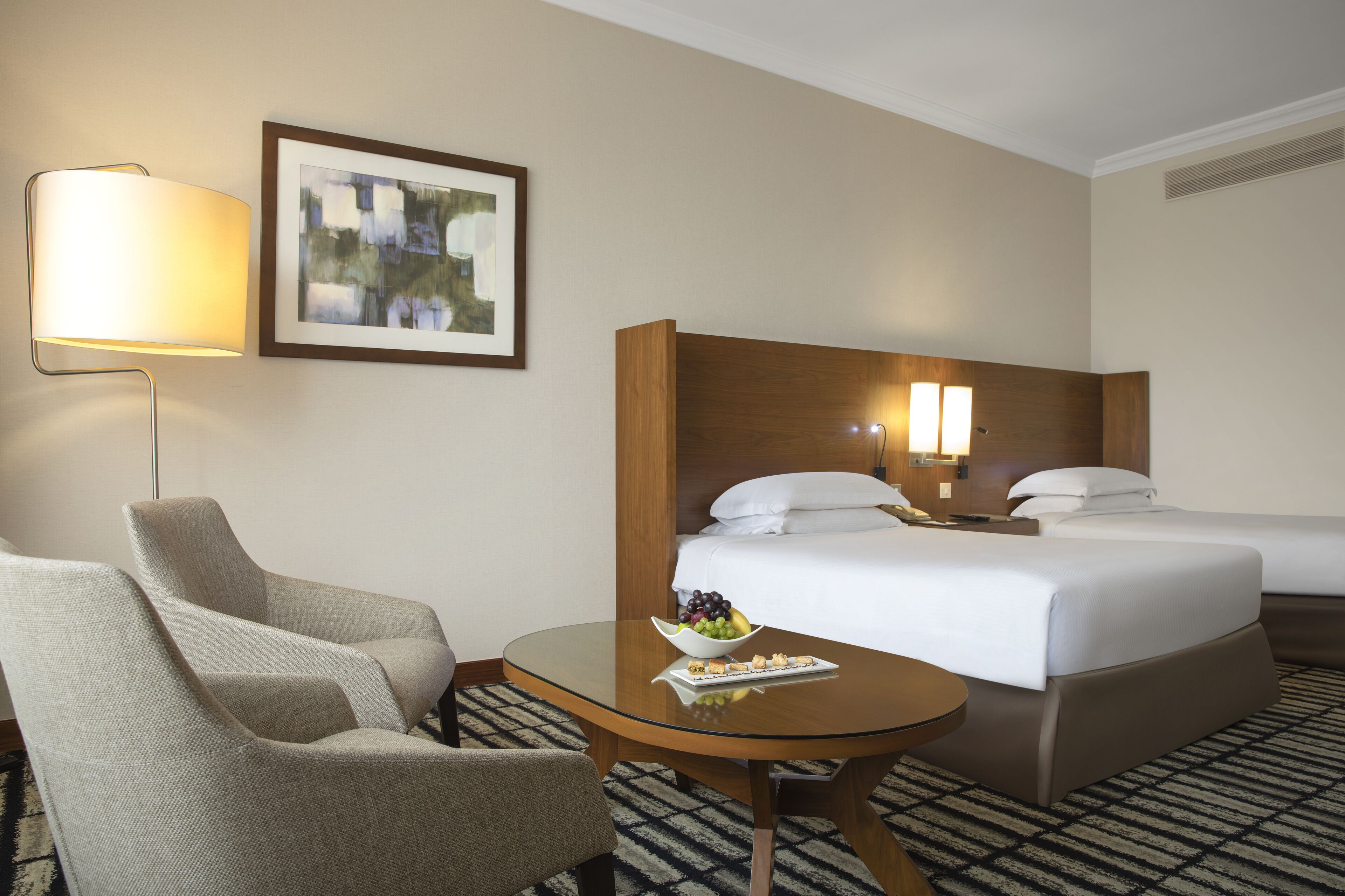 Ein Hotelzimmer mit zwei Betten, einem Sessel, einem Tisch, einer Lampe und einem gerahmten Bild an der Wand.