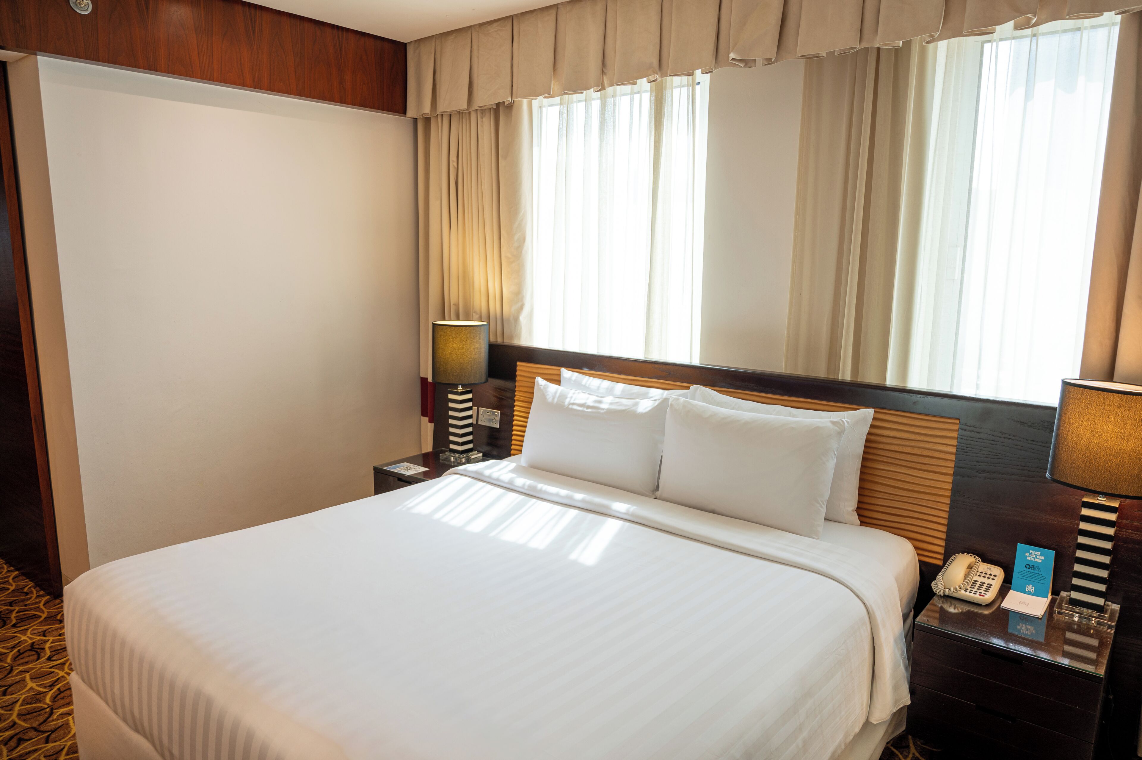 A hotel room with a bed, a nightstand with a lamp, a telephone, and a window with curtains.