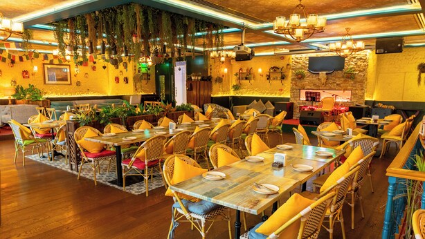 A restaurant with wicker chairs, wooden tables, hanging plants, and a stone wall.